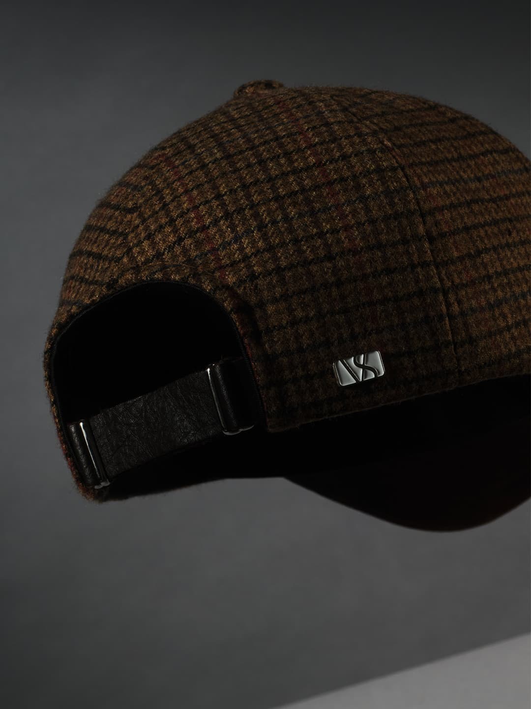 A brown plaid hat with a leather strap and metal buckle, partially in shadow against a gray background.