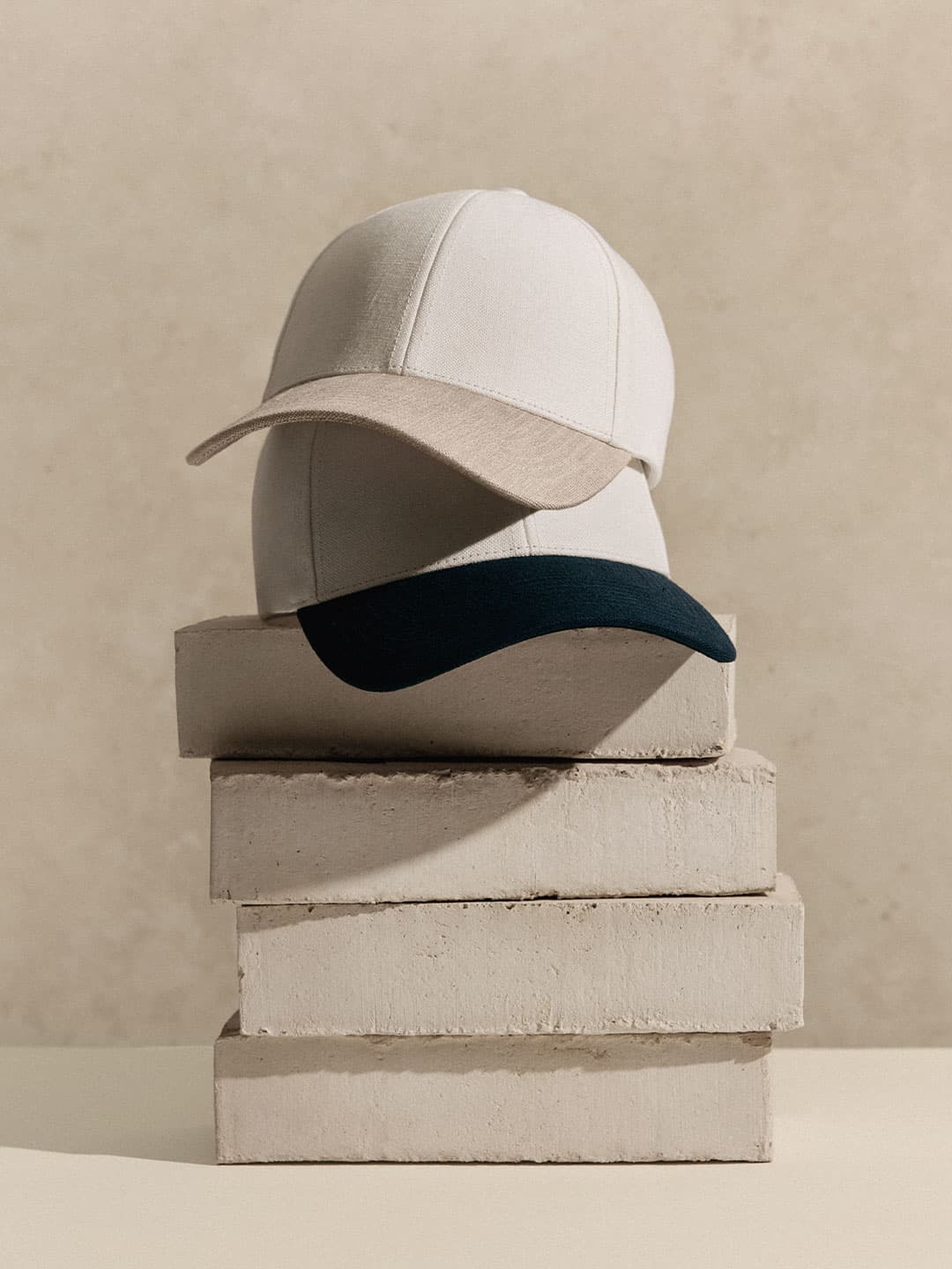 Two baseball caps, one with a beige and the other with a dark brim, stacked on concrete blocks against a neutral background.