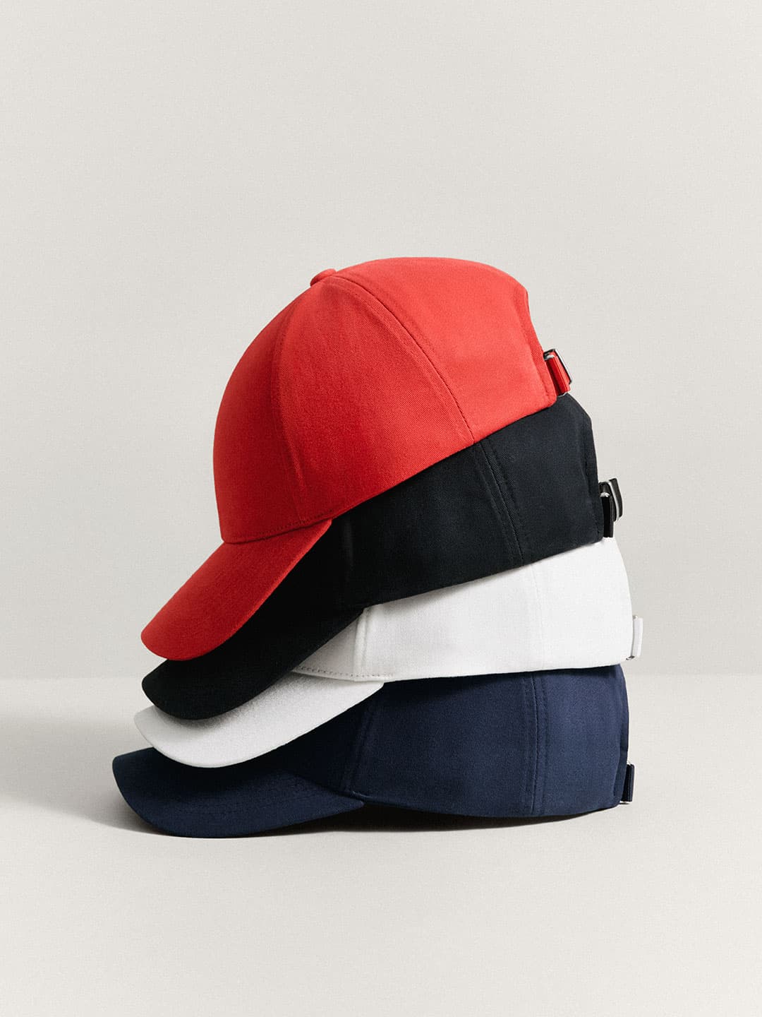 Stack of four baseball caps in red, black, white, and navy blue in Cotton Twill from Varsity Headwear, arranged neatly on a light surface against a neutral background.