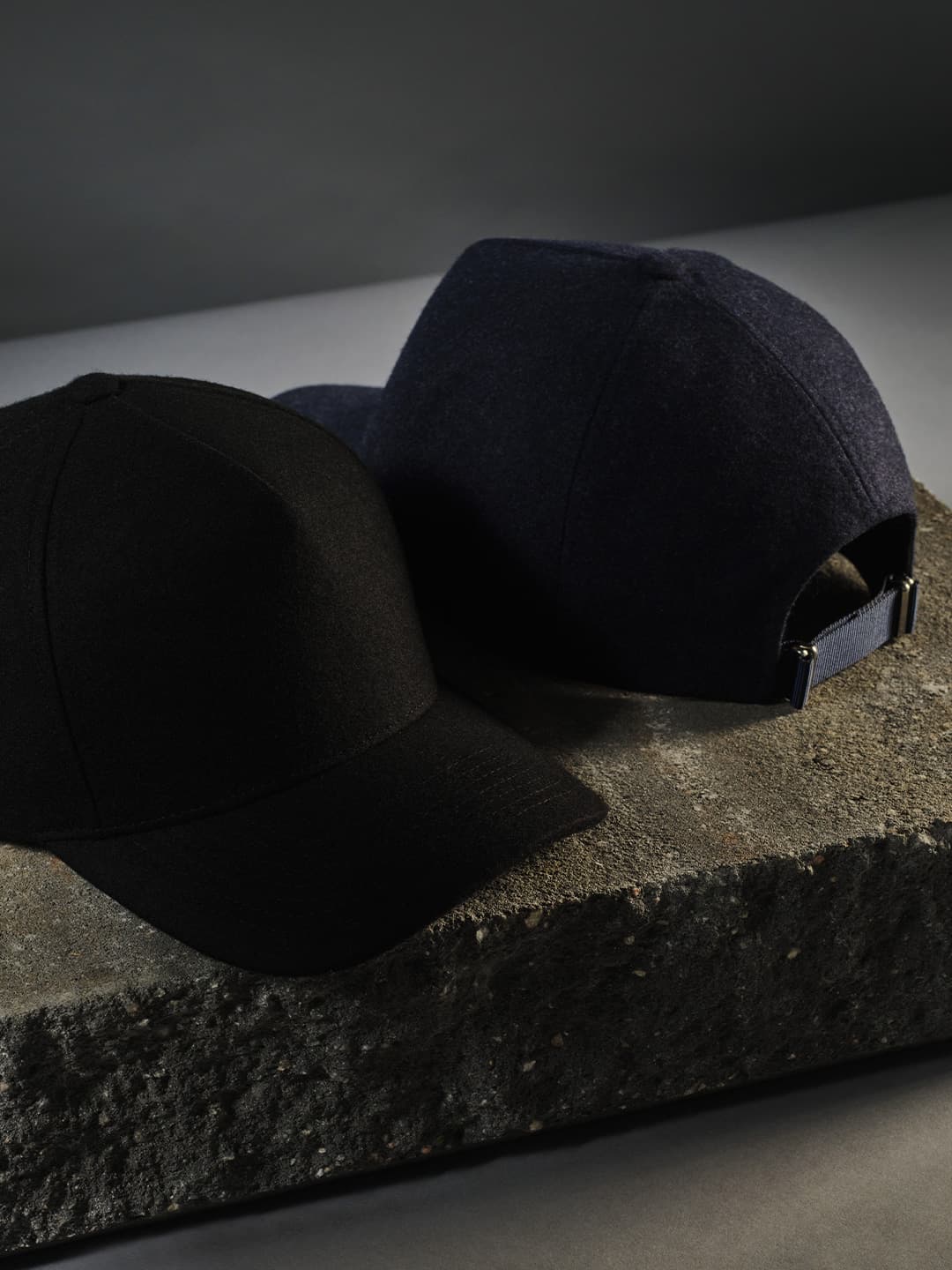 Two caps, one black and one navy, rest on a rough, textured concrete surface against a dark background.