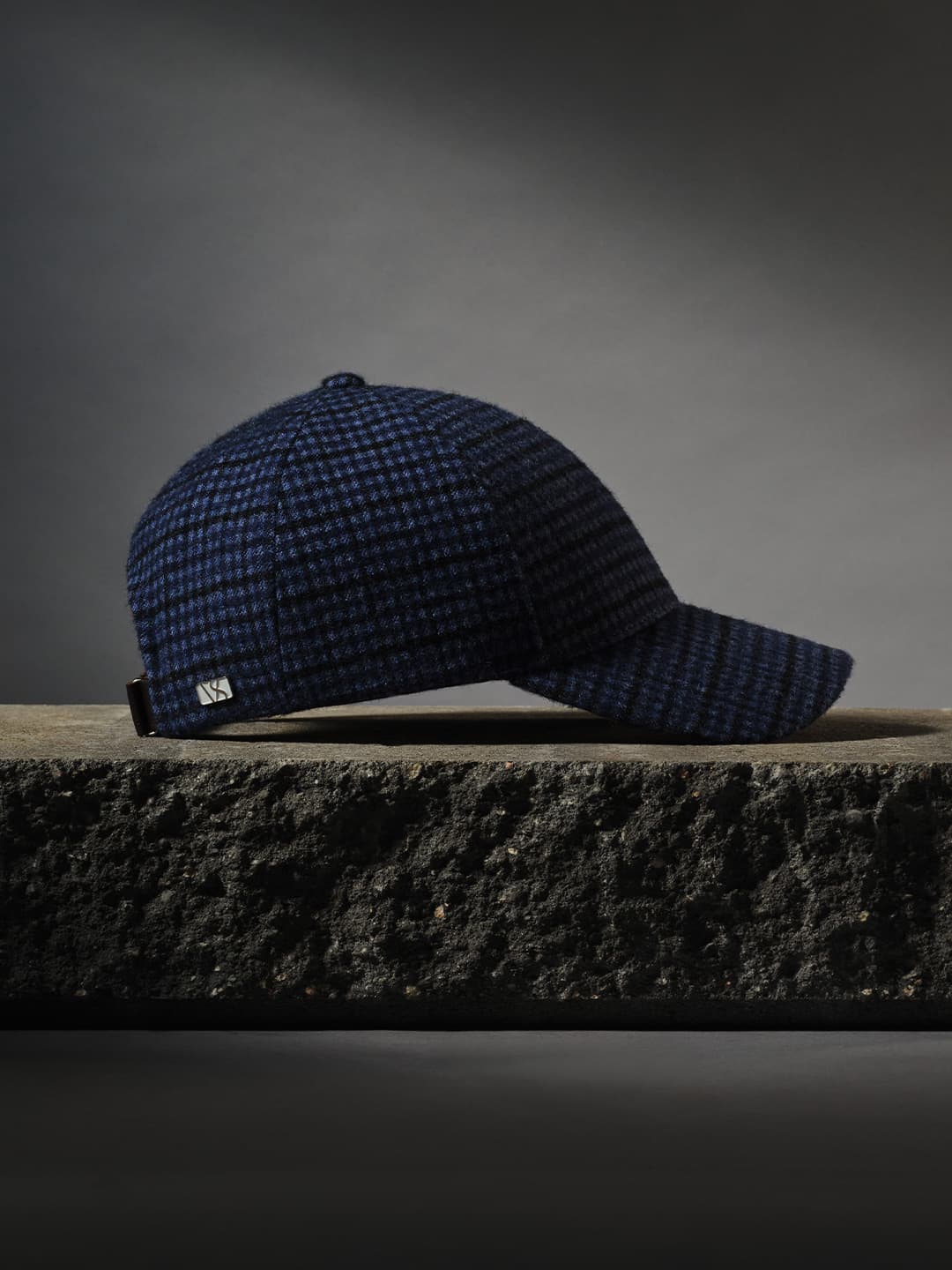 A blue checkered baseball cap with a curved brim rests on a textured black surface against a dark, neutral background.