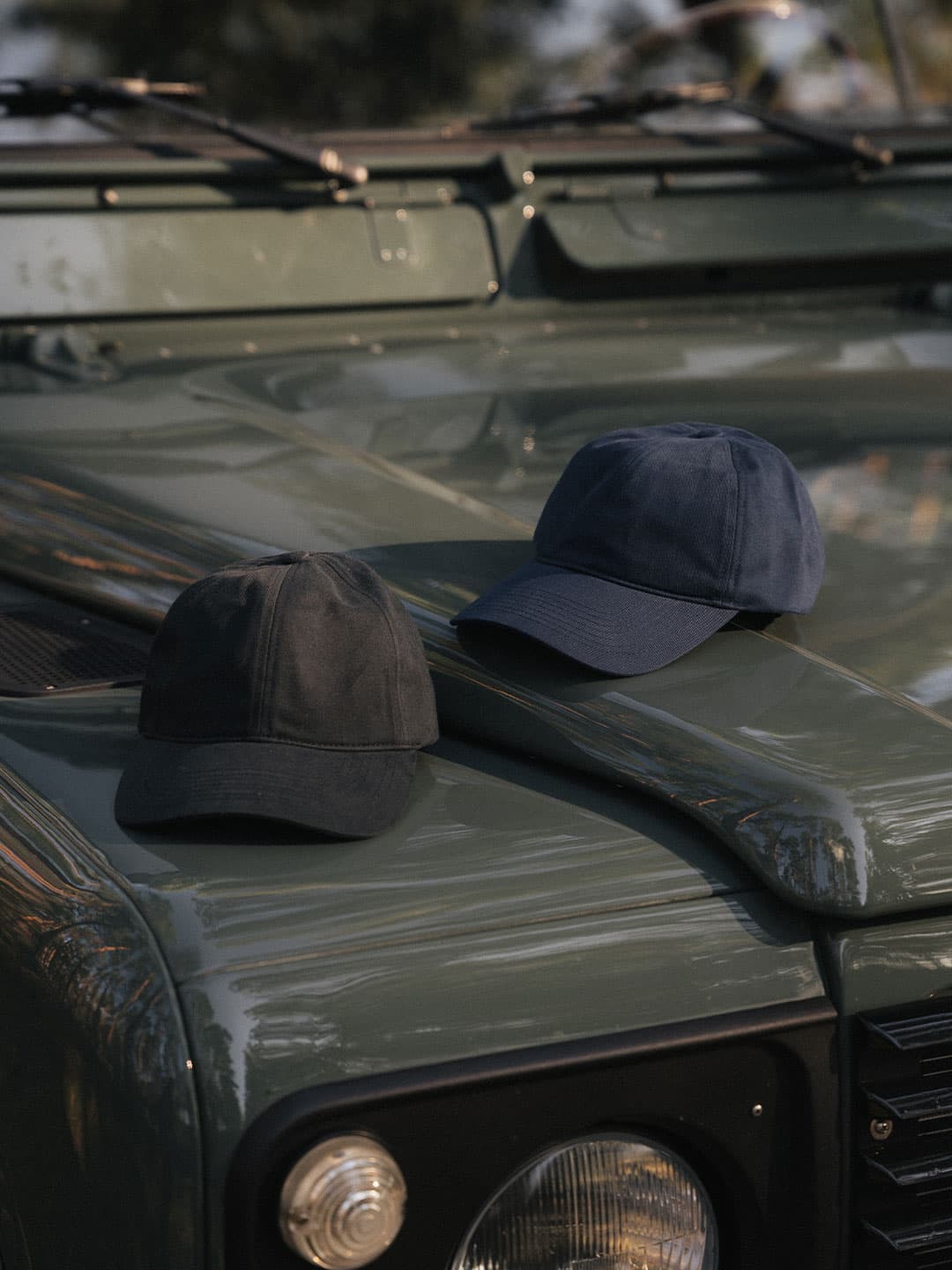 Two caps, one black and one blue, rest on the hood of a vintage green vehicle under soft daylight.
