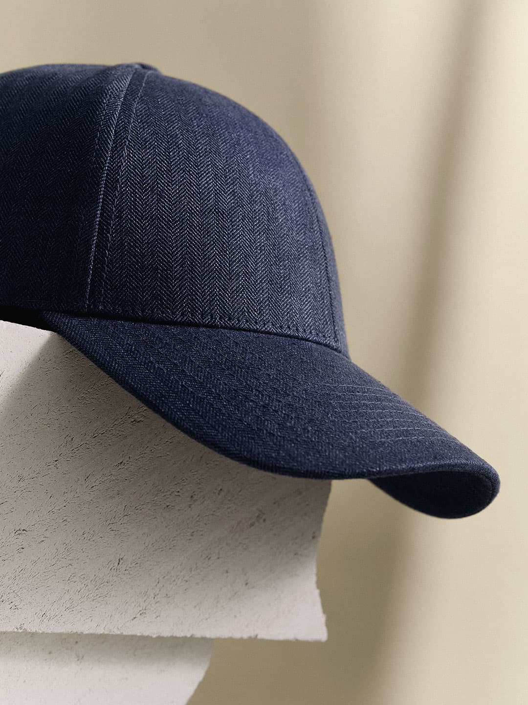 Close-up of a navy blue herringbone-patterned baseball cap resting on a textured white surface with a blurred beige background.