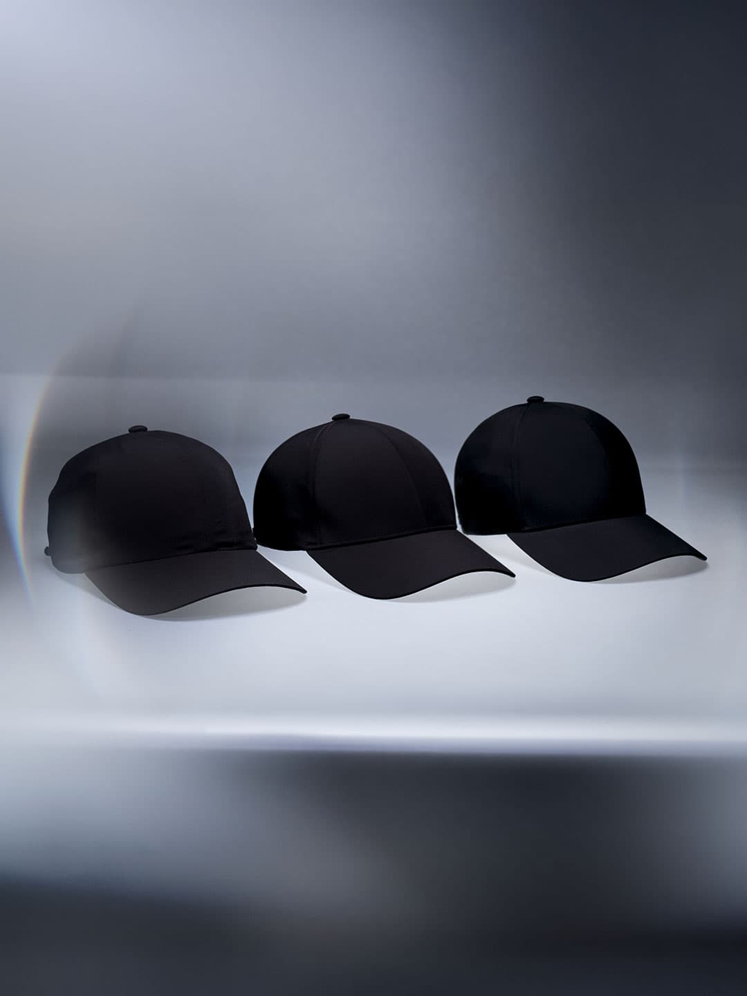 Three black sports caps lined up against a blurred, gradient background, each with a slightly different design.