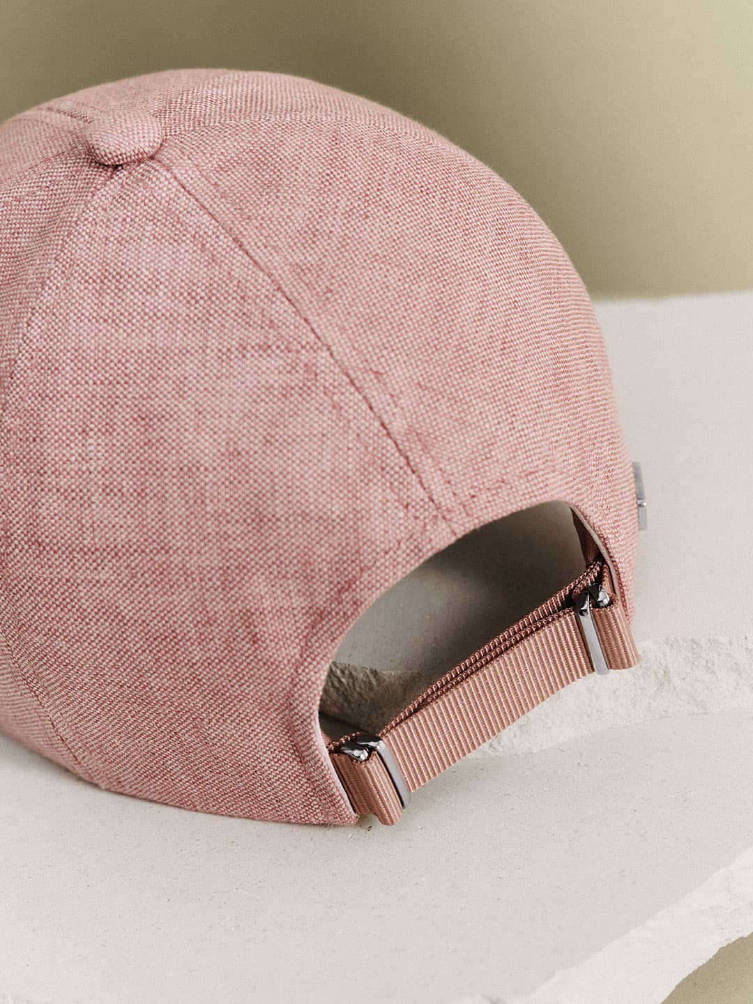 Close-up of the back of a pink fabric baseball cap with an adjustable strap, resting on a white textured surface.