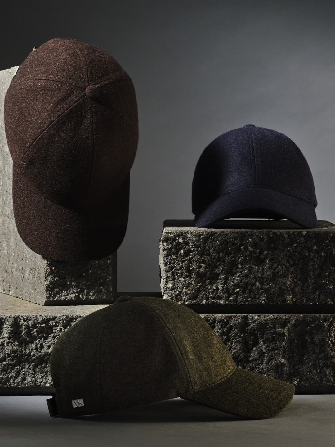 Three textured baseball caps in brown, navy, and olive are displayed on rugged stone blocks against a dark background.