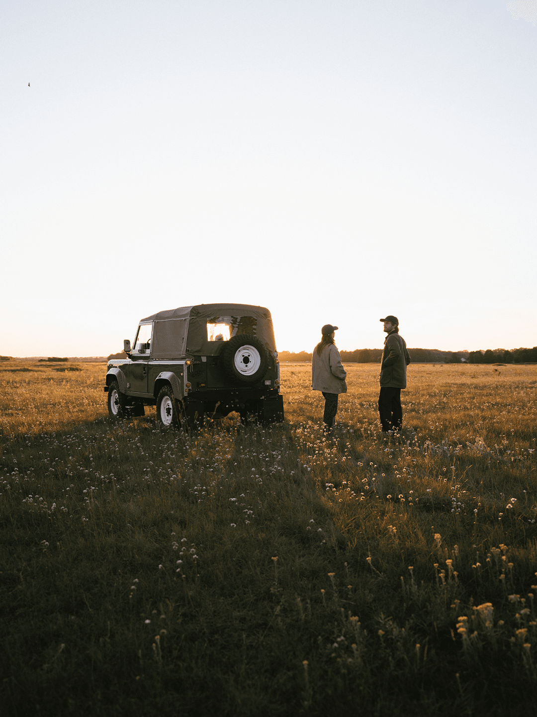 Two people stand near a parked off-road vehicle in a grassy field at sunset, engaged in conversation.