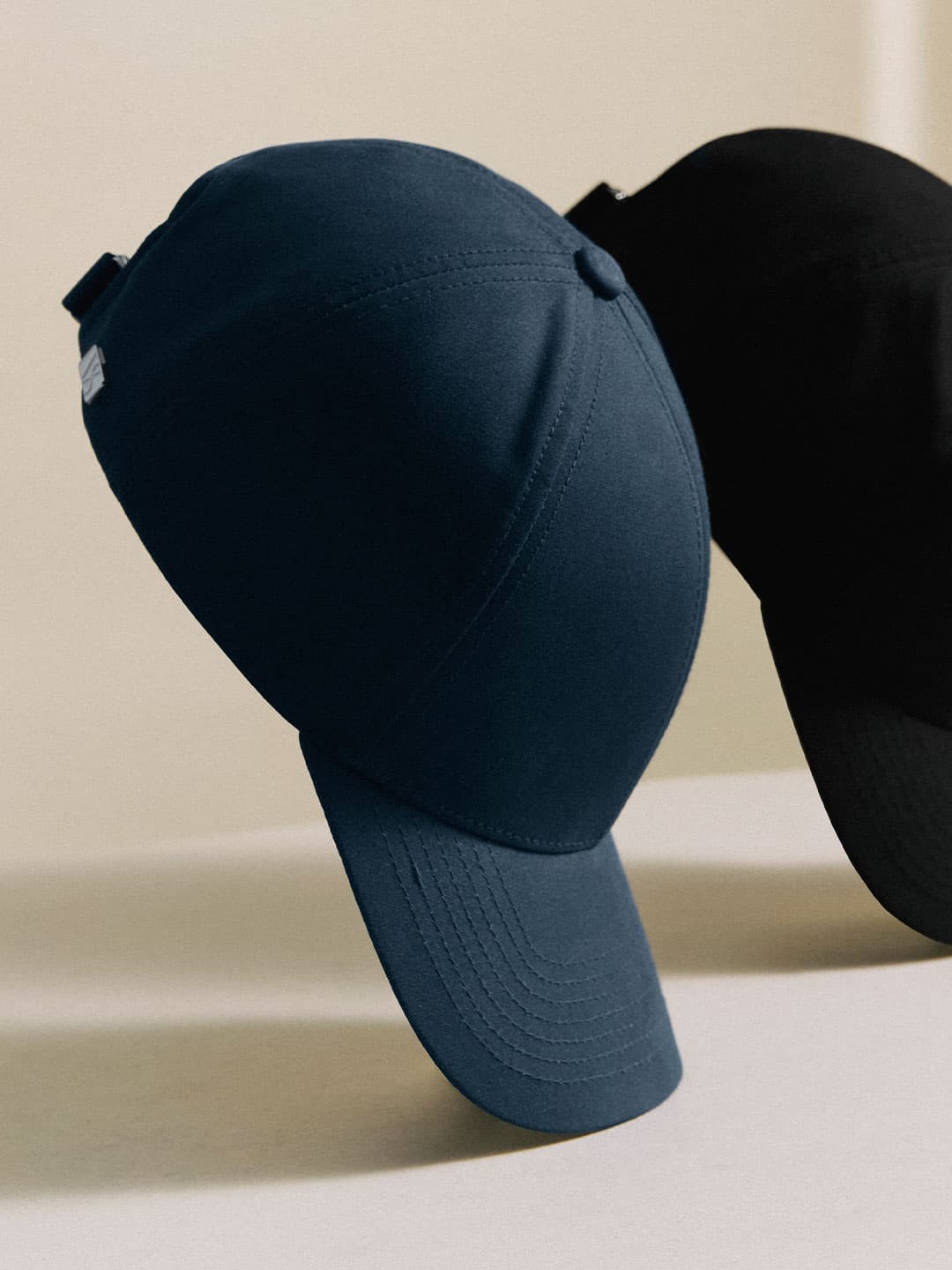 Two baseball caps, one navy and one black, are positioned upright on a light surface with a neutral background.