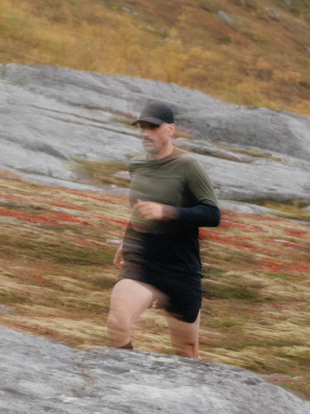 A man in a cap and athletic wear running on a rocky terrain with autumn foliage in the background, captured with motion blur.