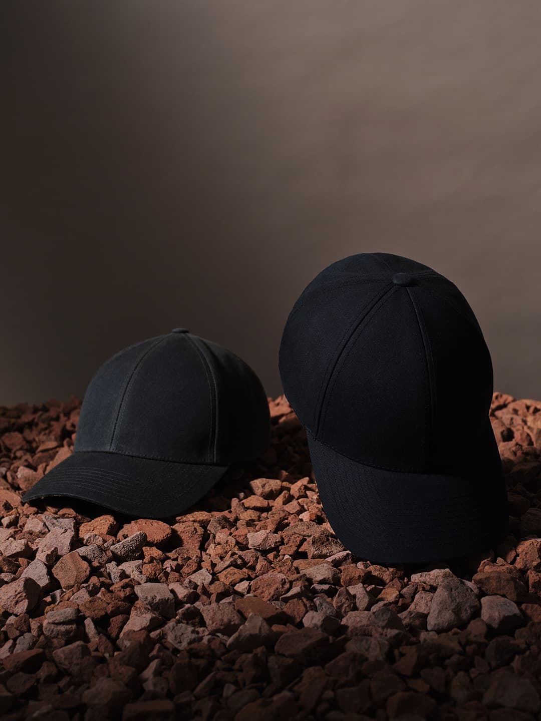 Two black baseball caps on a bed of reddish-brown stones, with a neutral gradient background.