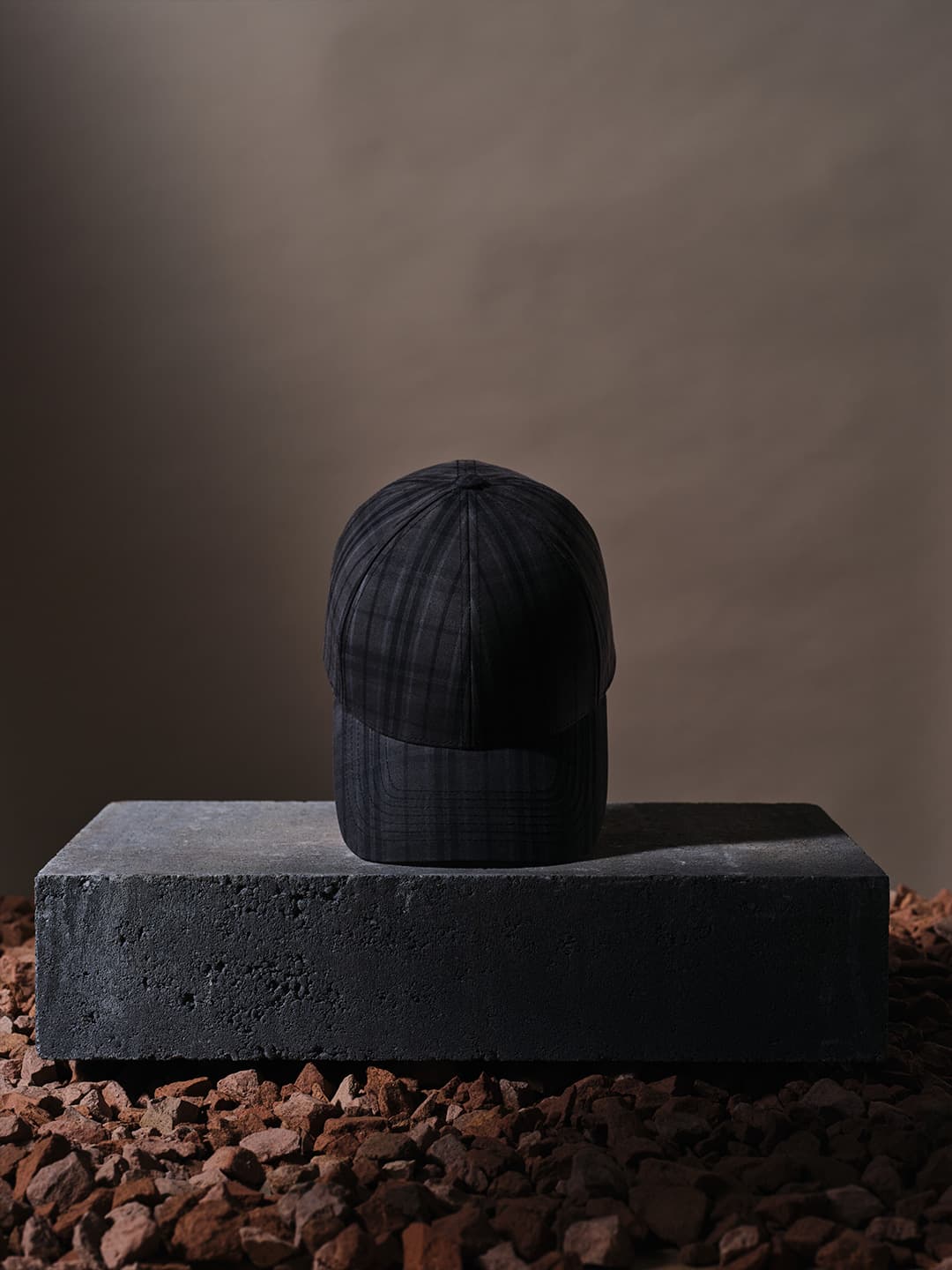 A black plaid cap sits on a concrete block surrounded by reddish-brown stones, against a plain brown background.