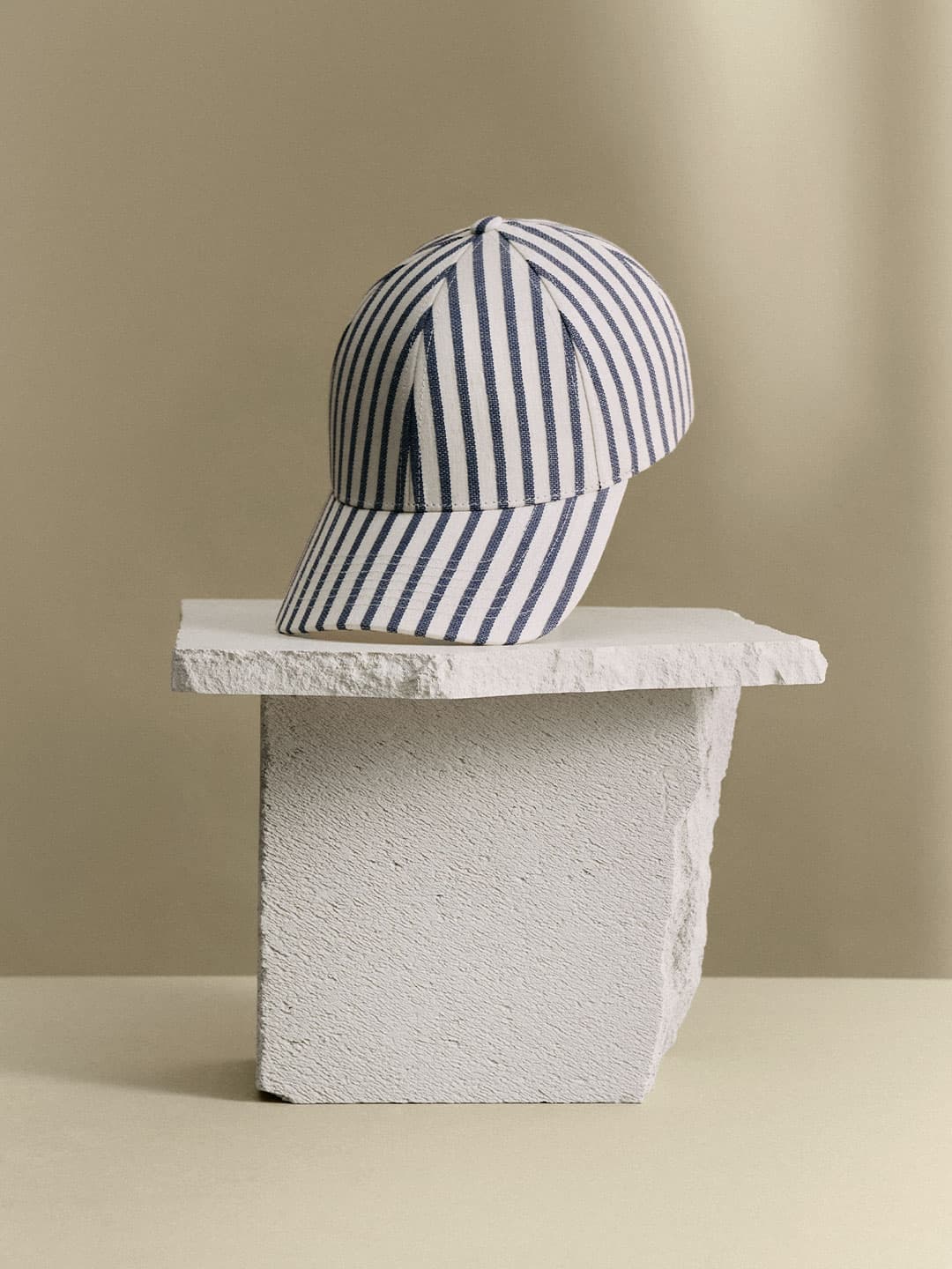 Striped baseball cap with blue and white vertical lines displayed on a textured rectangular stone block against a beige background.