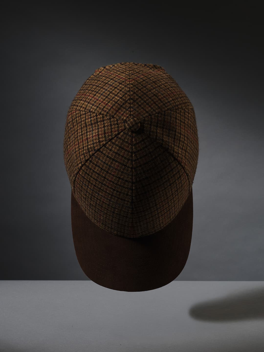 A beige and brown plaid cap with a solid brown brim, suspended against a dark, moody background.