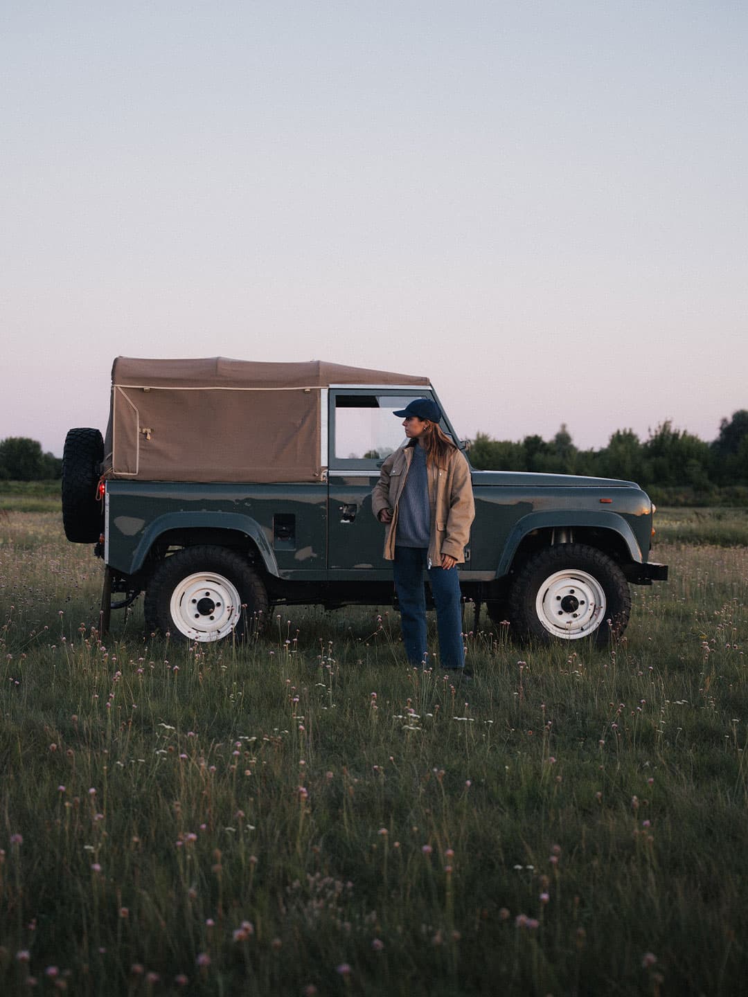 Person standing beside a vintage SUV in a field at dusk, wearing a varsity headwear cotton twill cap and jacket, with the sky softly lit in the background.