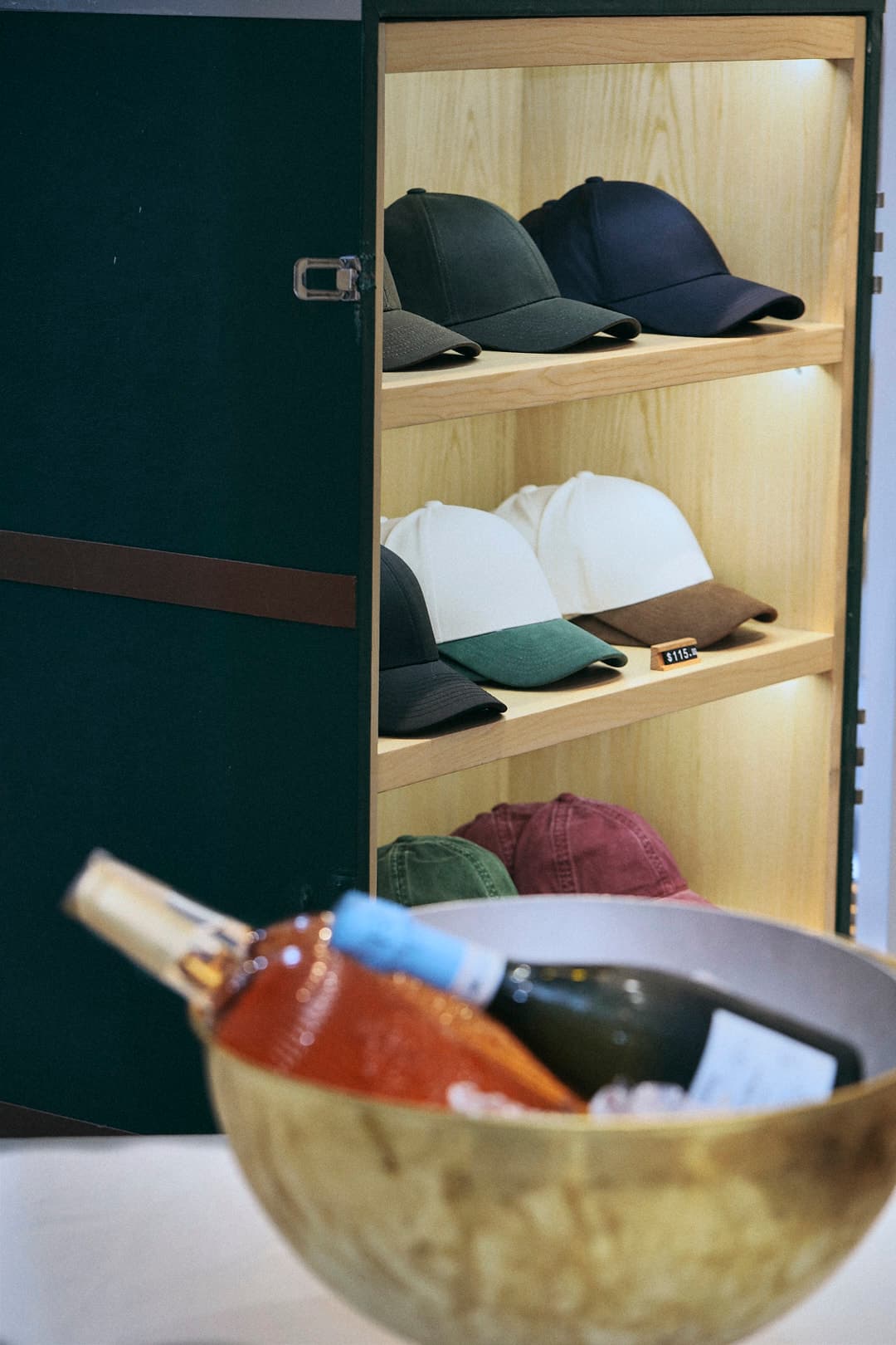A wooden shelf displaying various colored baseball caps inside a cabinet, with a bowl of wine bottles in the foreground.