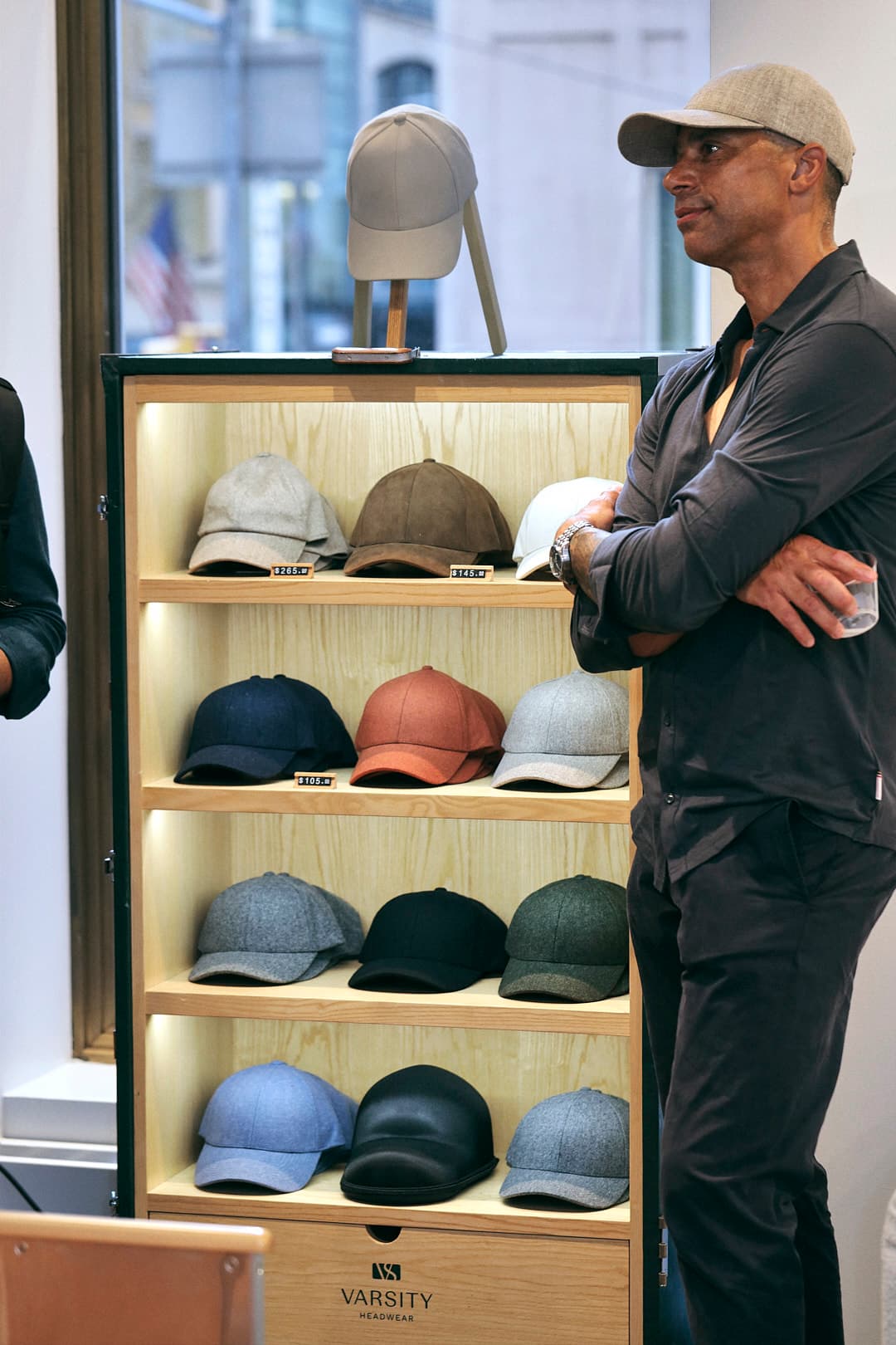 A man wearing a cap stands beside a display of various colored caps on shelves inside a store.