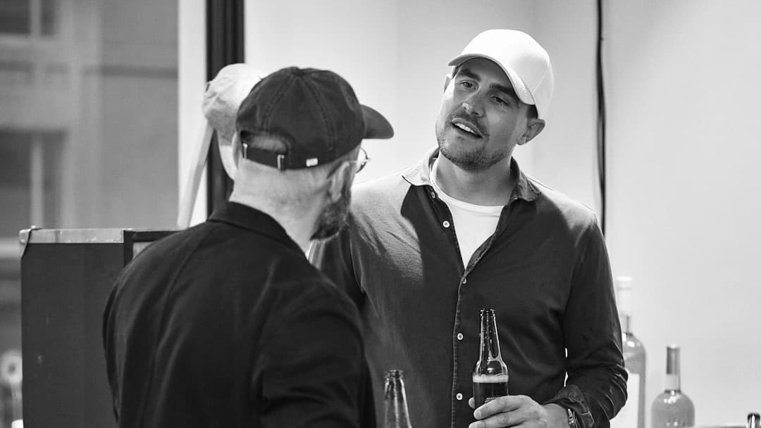 Two men wearing hats engage in conversation at a casual gathering, one holding a beer bottle.
