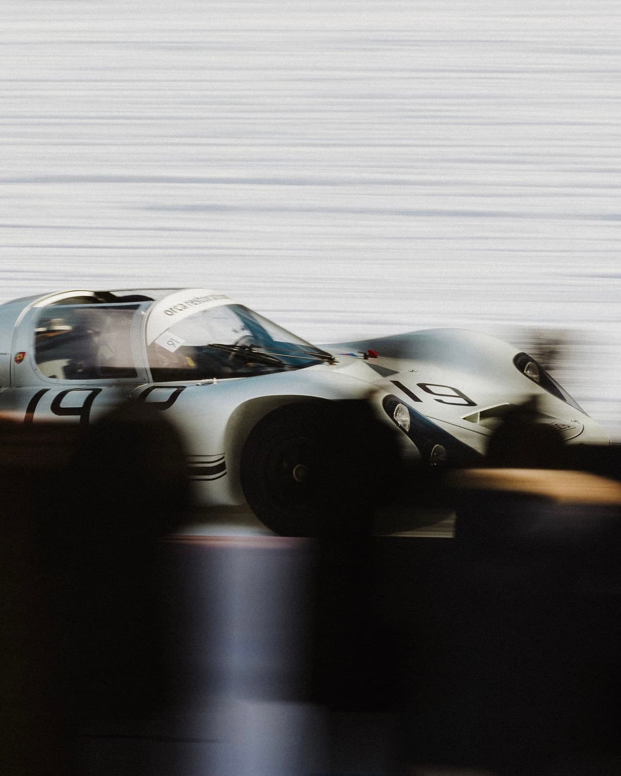 Vintage white race car with the number 19 on the side, speeding on a track, with blurred motion in the foreground and background.