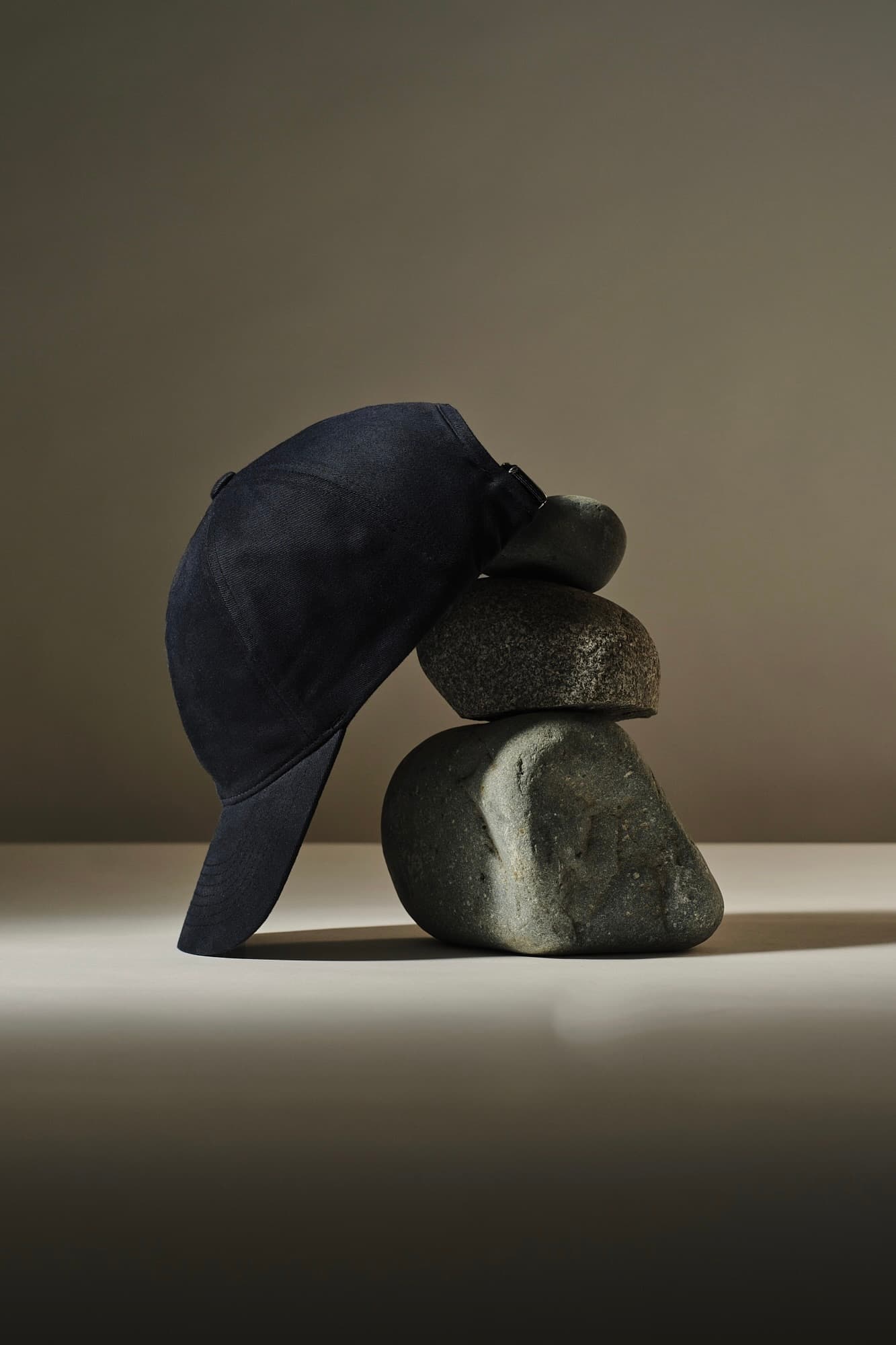 A dark baseball cap rests on a stack of three smooth, gray stones in a balanced arrangement against a neutral background.