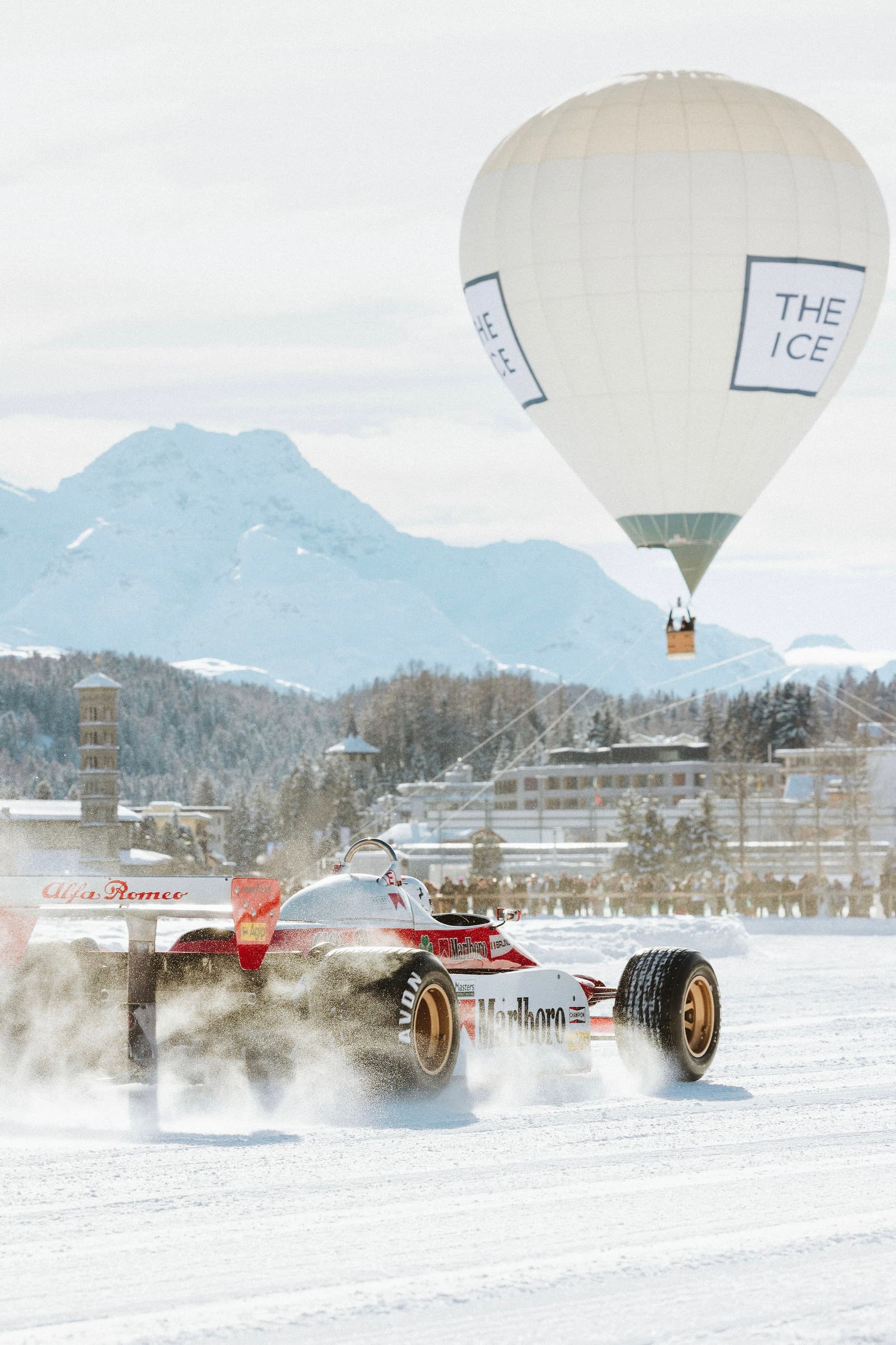 A vintage race car speeds on snow with a large hot air balloon in the sky, at The I.C.E in St. Moritz