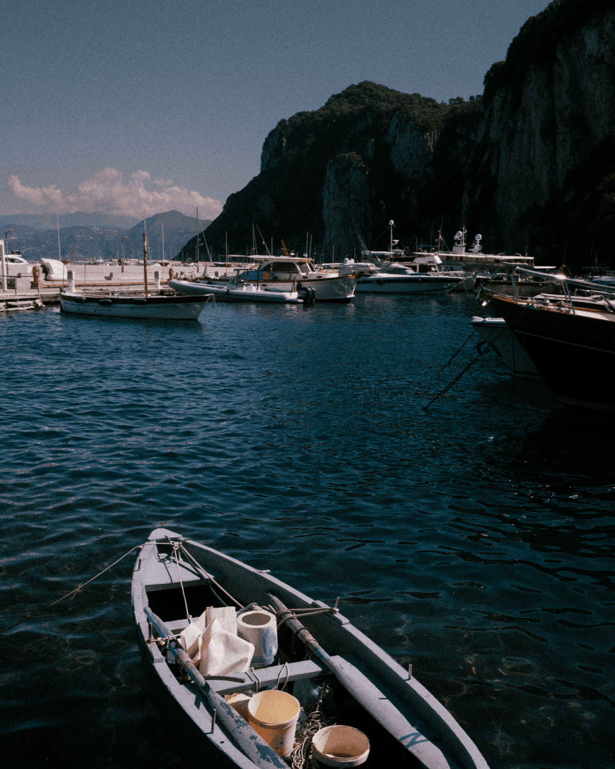 A small rowboat with supplies floats in a marina, surrounded by yachts and steep cliffs under a clear blue sky.