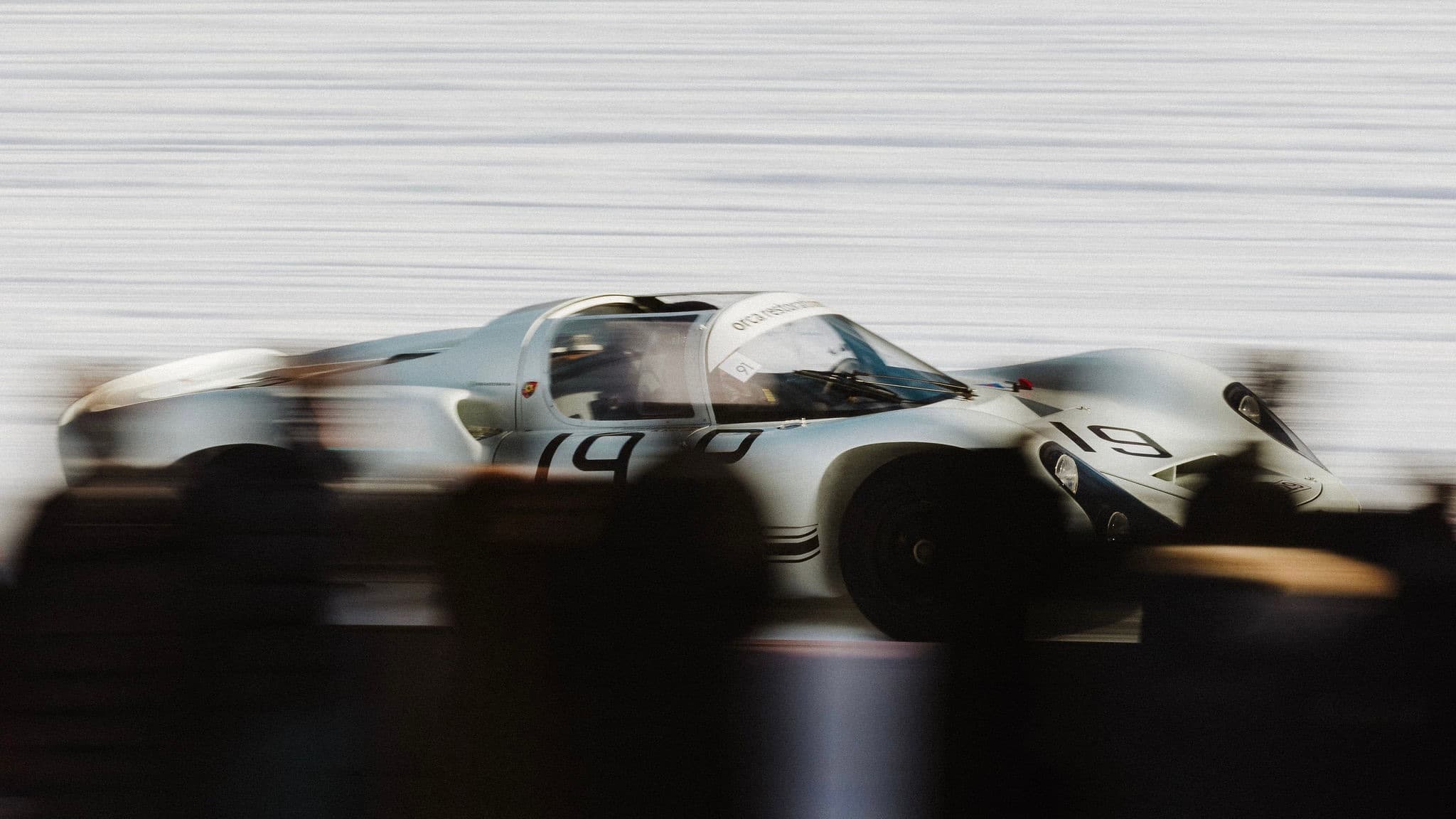 A vintage silver race car speeds past blurred spectators on a track, with the number 19 visible on its side at The I.C.E in St. Moritz