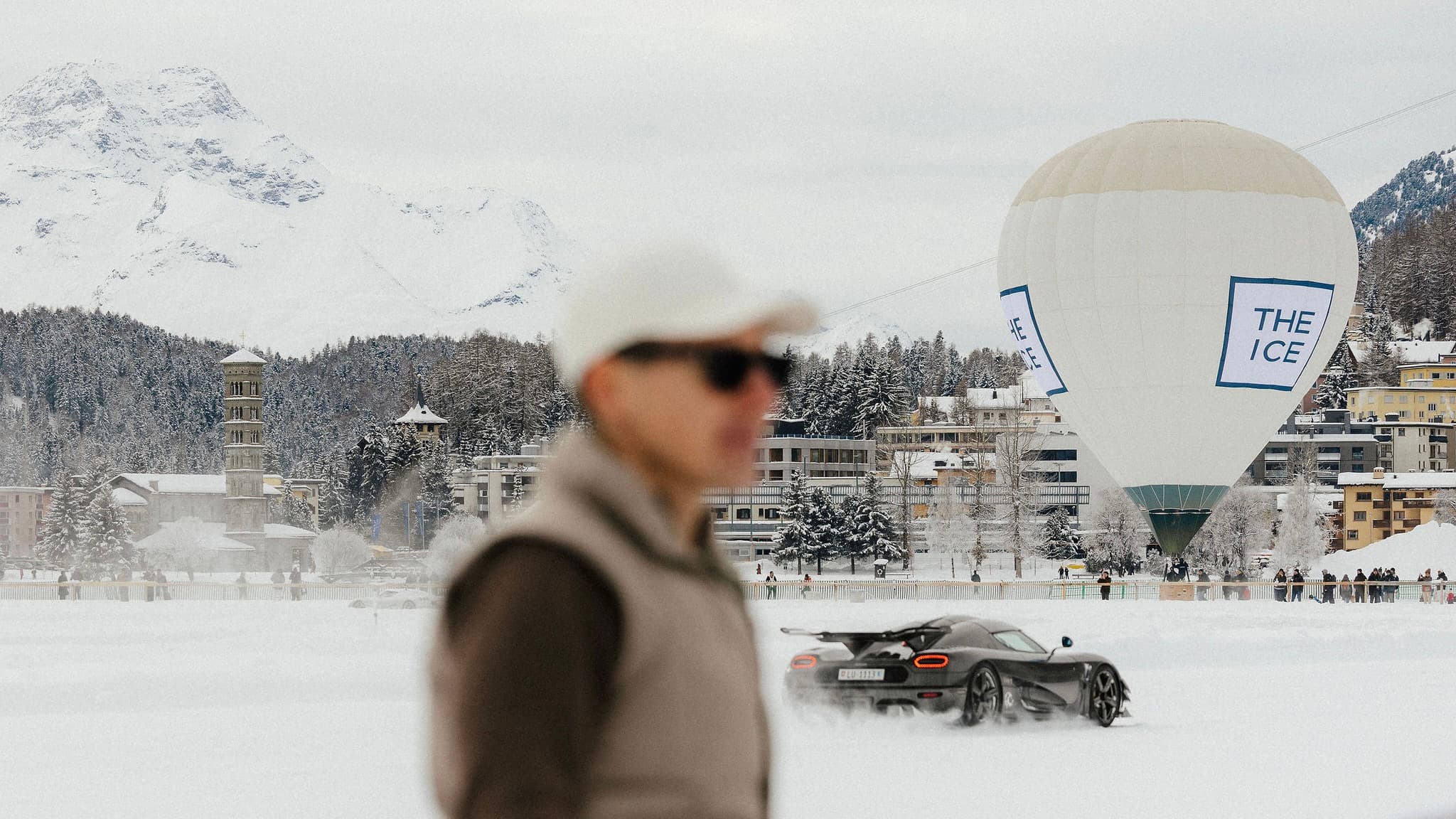 Photographer Joel Hyppönenin stands with a snowy landscape, a sports car drifting, and a hot air balloon in the background at The I.C.E St. Moritz
