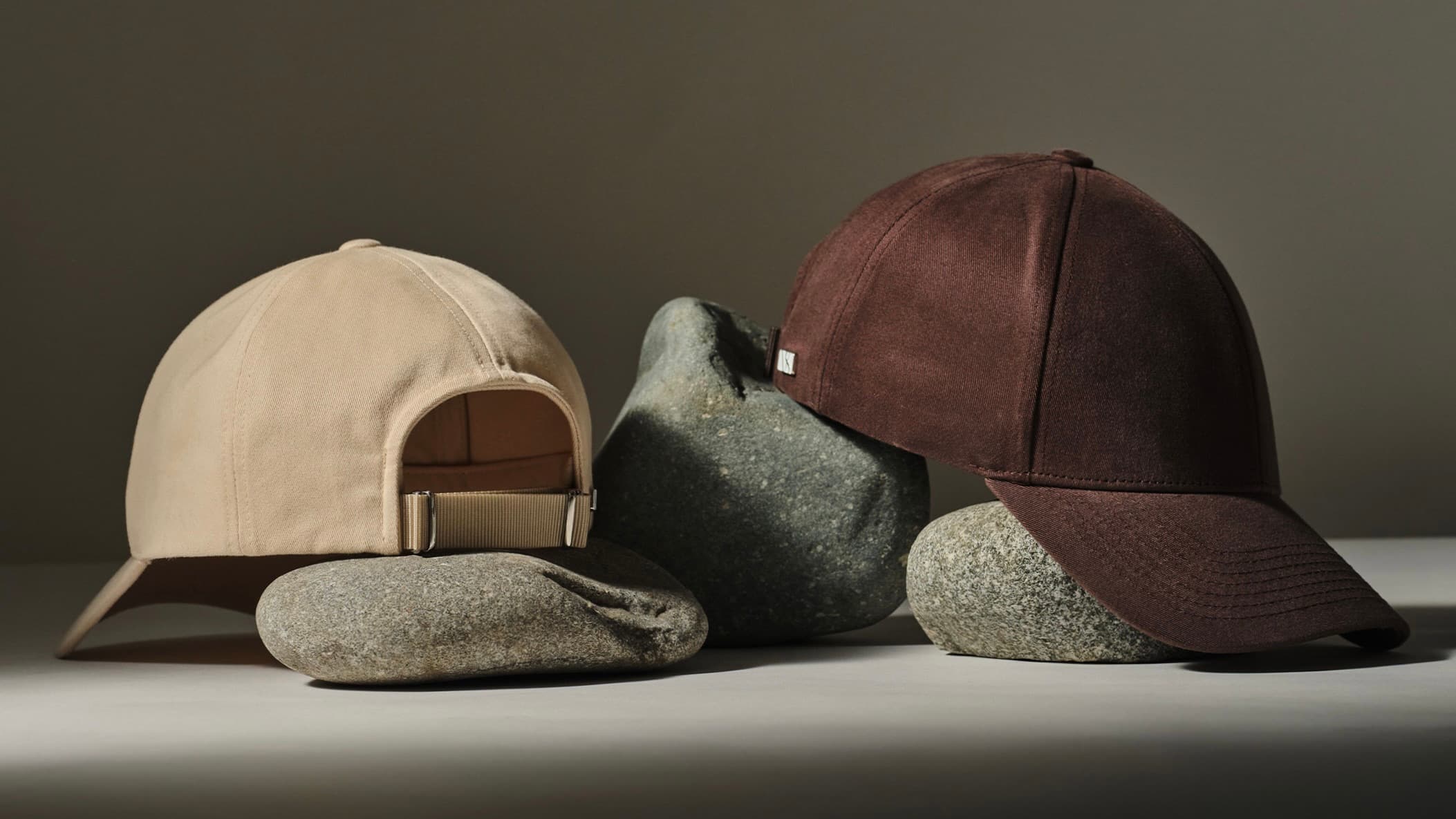 Two baseball caps, one beige and one brown, are creatively displayed on smooth grey stones against a neutral background.