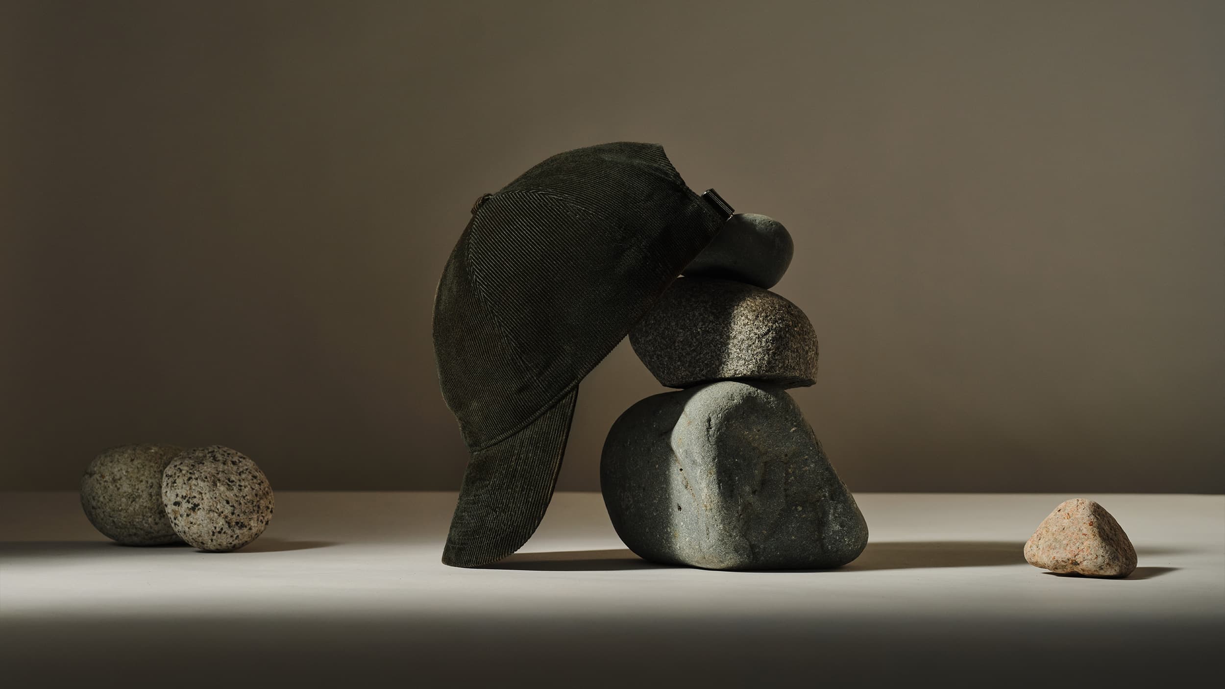 A dark green baseball cap balanced on a stack of smooth gray stones with additional stones surrounding it, set against a neutral background.