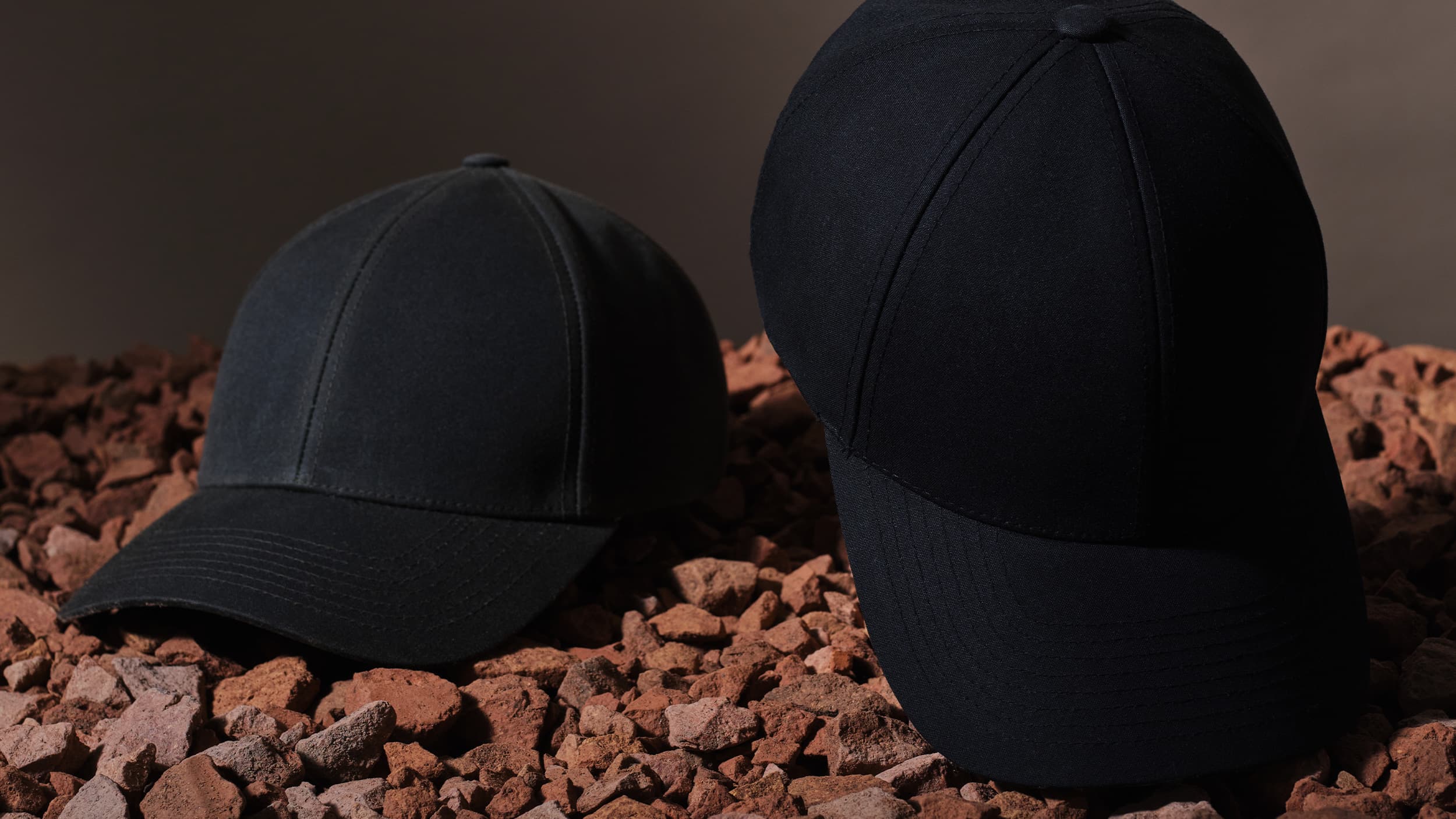 Two black baseball caps on reddish-brown pebbles, dimly lit with a neutral background.