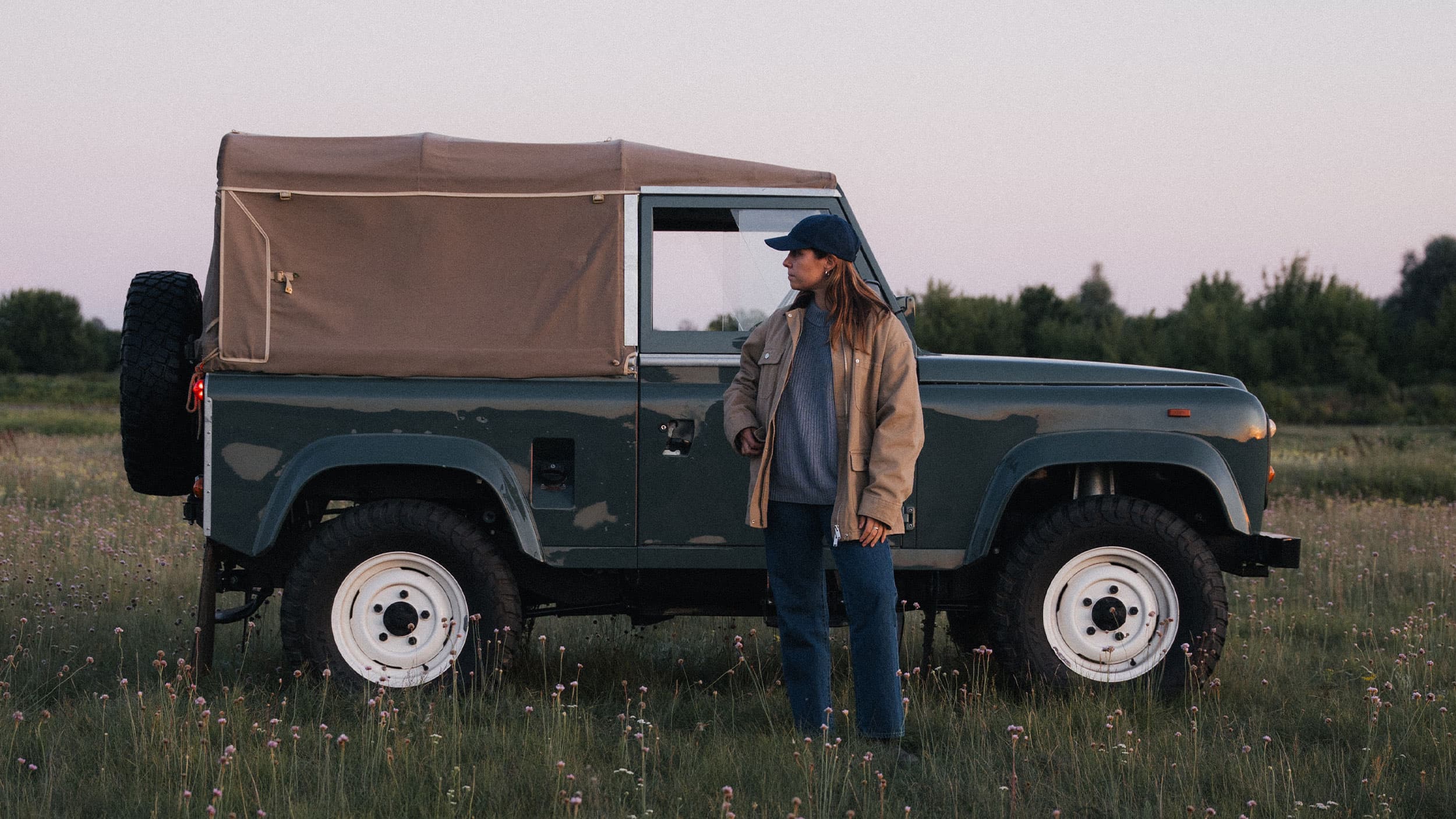 A person in a jacket and cotton twill varsity headwear cap stands beside a vintage off-road vehicle with a canvas top in a grassy field at dusk.