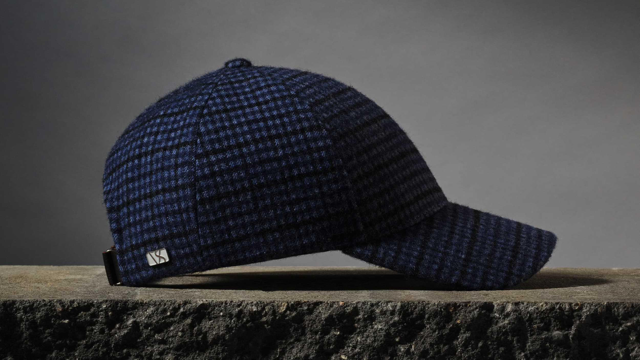 Dark blue checkered wool baseball cap with a small metal logo, resting on a textured stone surface against a gray background.
