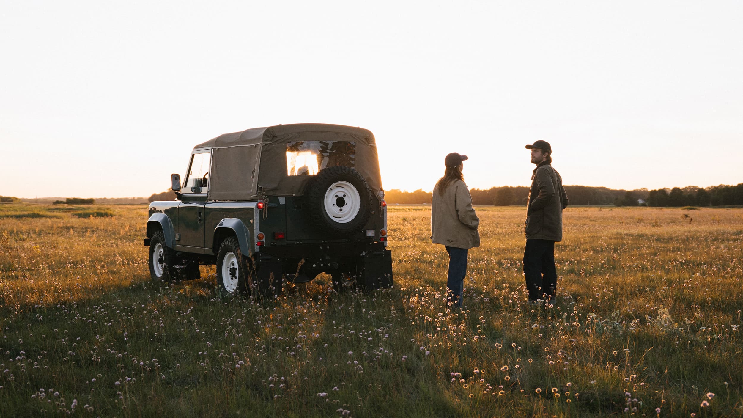 Two people stand beside a vintage SUV in a vast field during sunset, surrounded by tall grass and wildflowers.