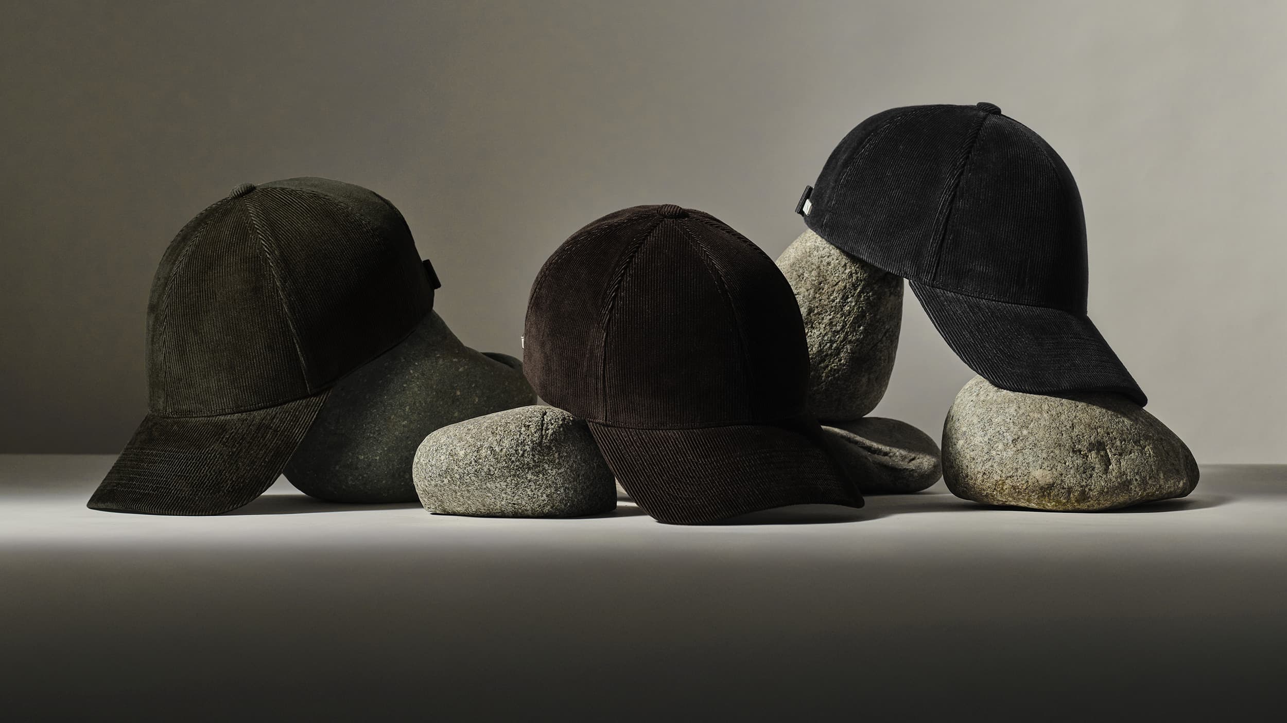 Three dark-colored baseball caps made of corduroy, displayed on smooth, rounded stones in a minimalist setting.