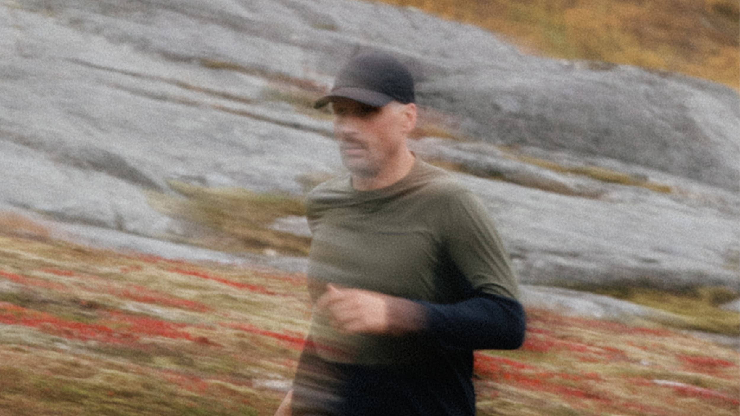 A man in a cap and green shirt jogging on a blurred natural landscape with rocks and grass.
