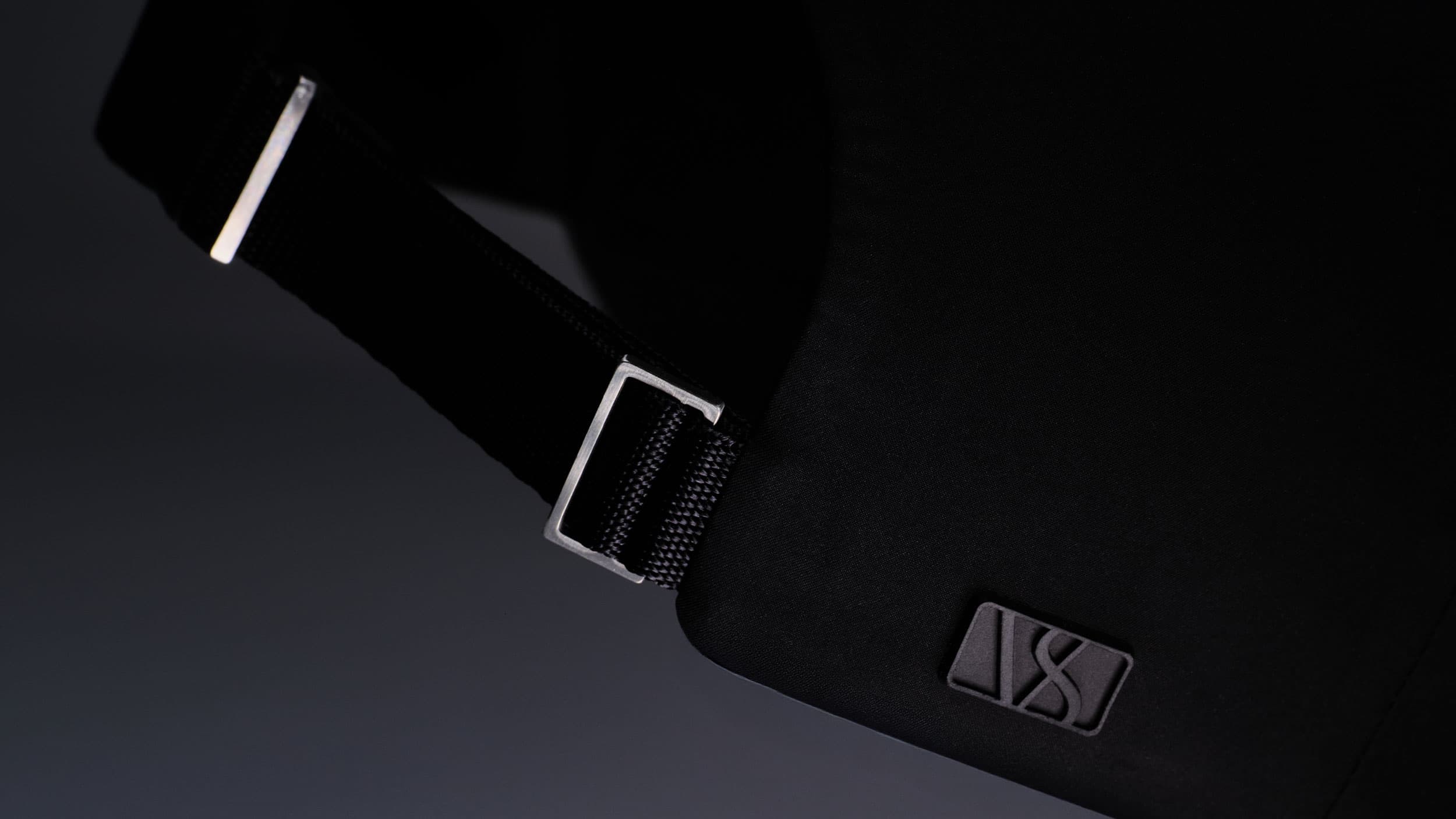 Close-up of a black baseball cap with a logo, featuring a strap and metallic buckle against a dark background.