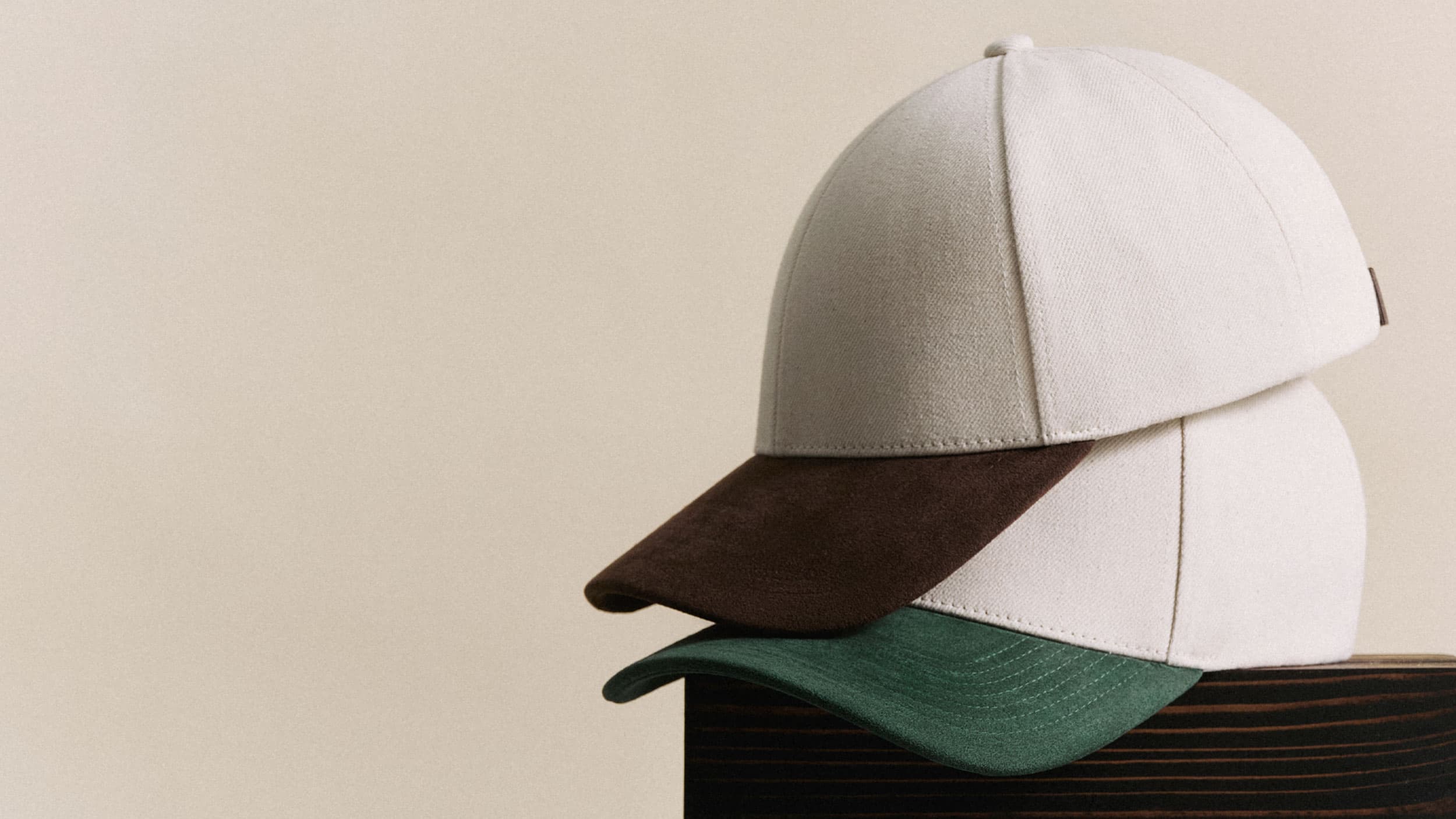 Stacked beige baseball caps with contrasting brown and green brims, placed on a wooden surface against a neutral background.