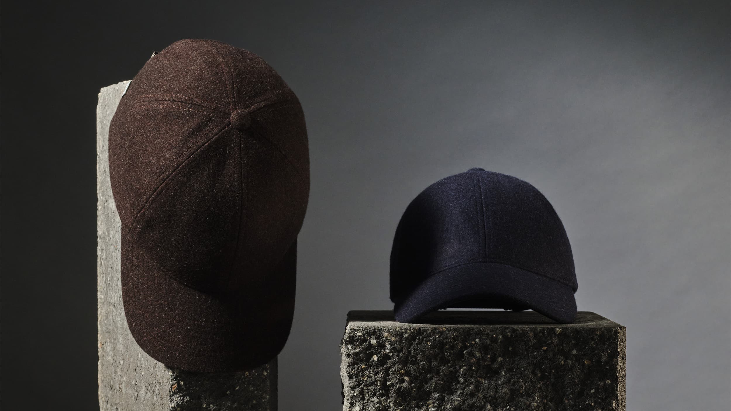 Two merino wool baseball caps, one brown and one navy, displayed on gray concrete pedestals against a gradient background.