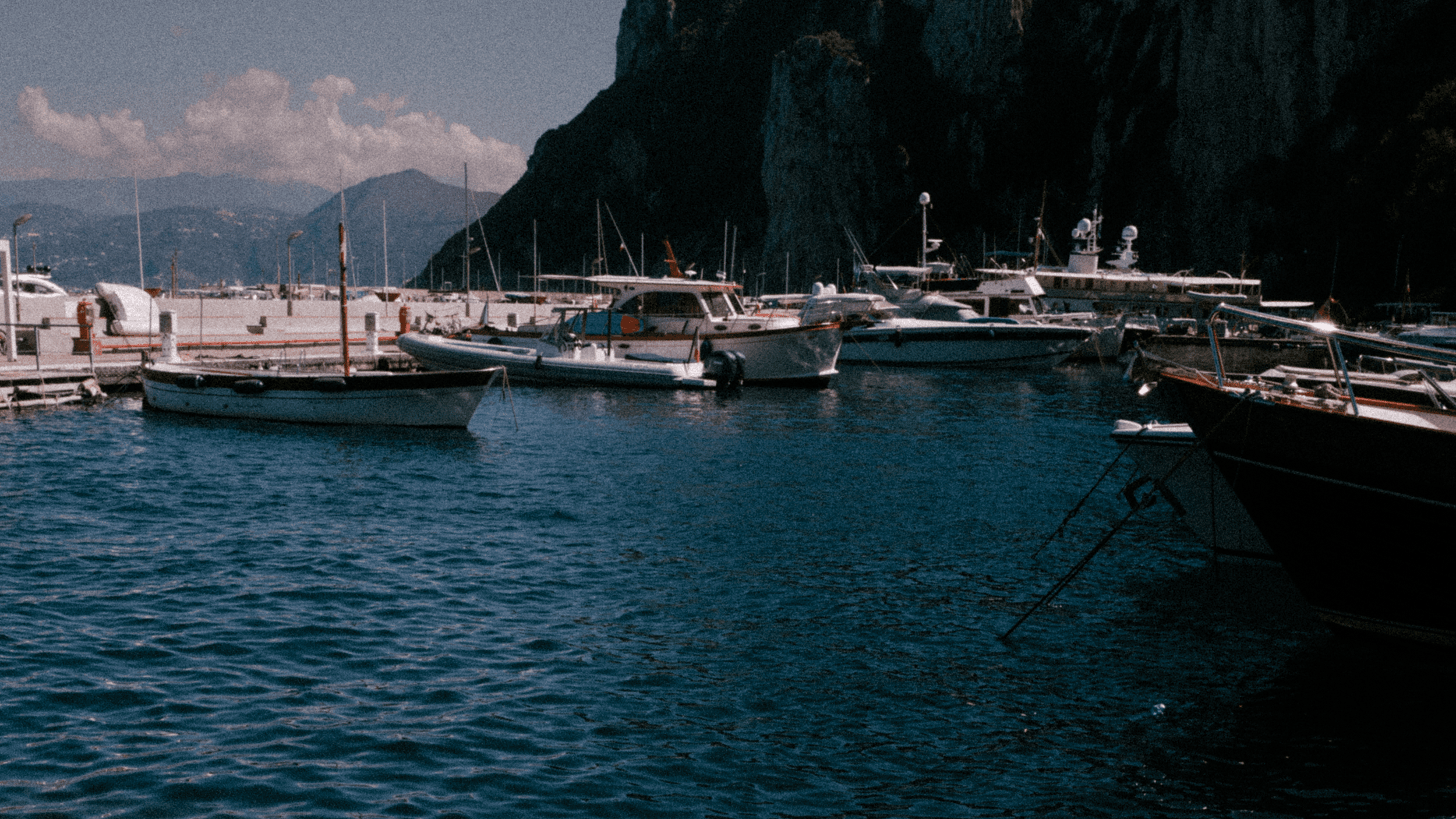 Boats docked at a marina with calm blue water, mountains, and cliffs in the background under a partly cloudy sky.