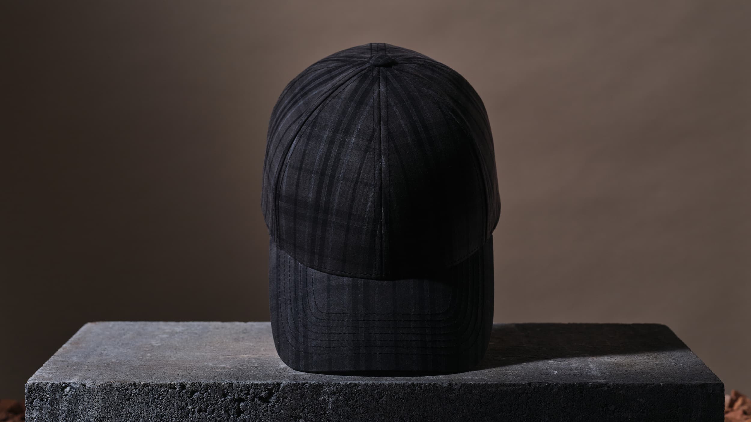 A black plaid baseball cap displayed on a textured stone pedestal against a neutral brown background.