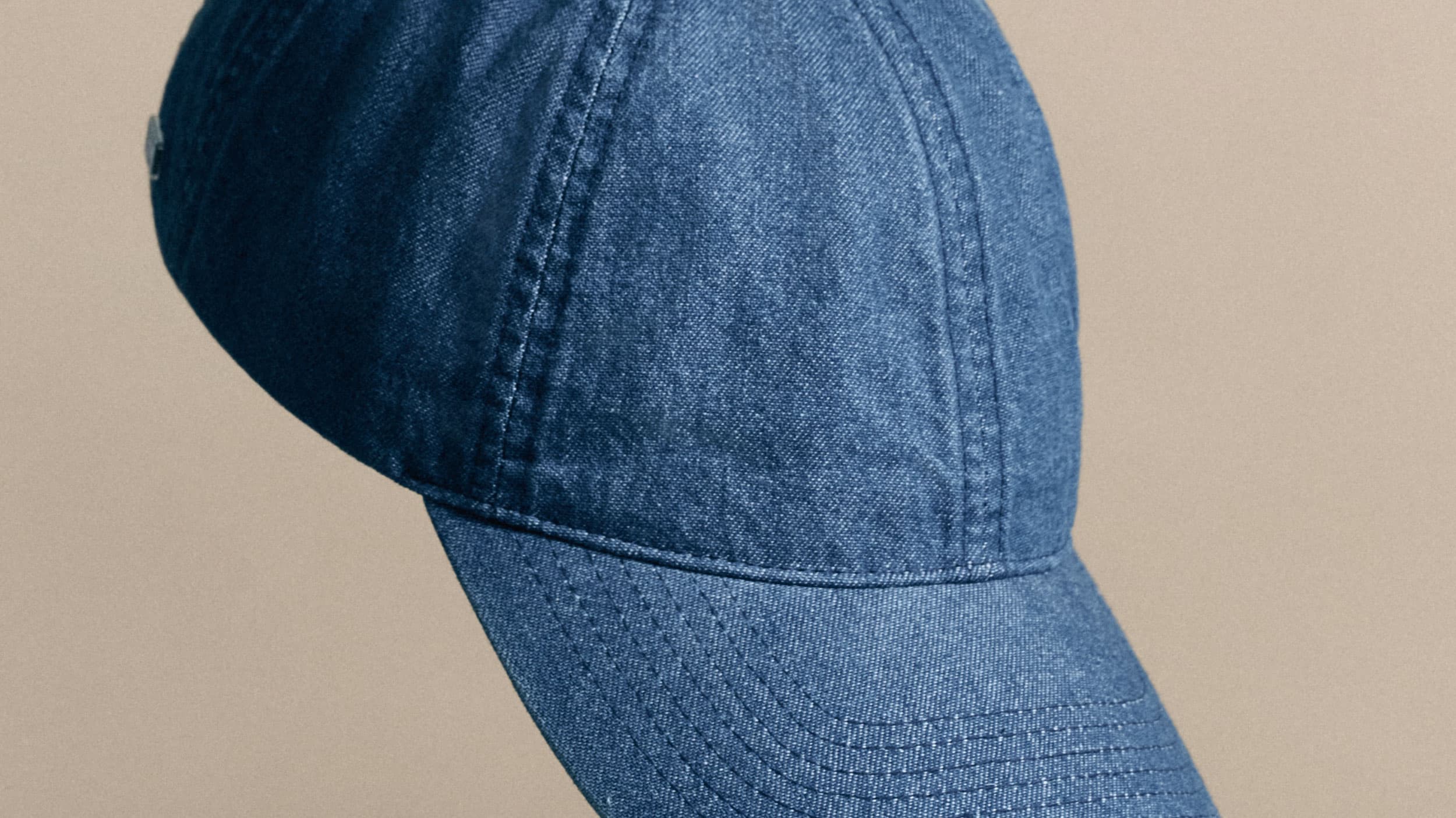 Close-up of a blue denim baseball cap against a neutral beige background. The cap features visible stitching and a curved brim.