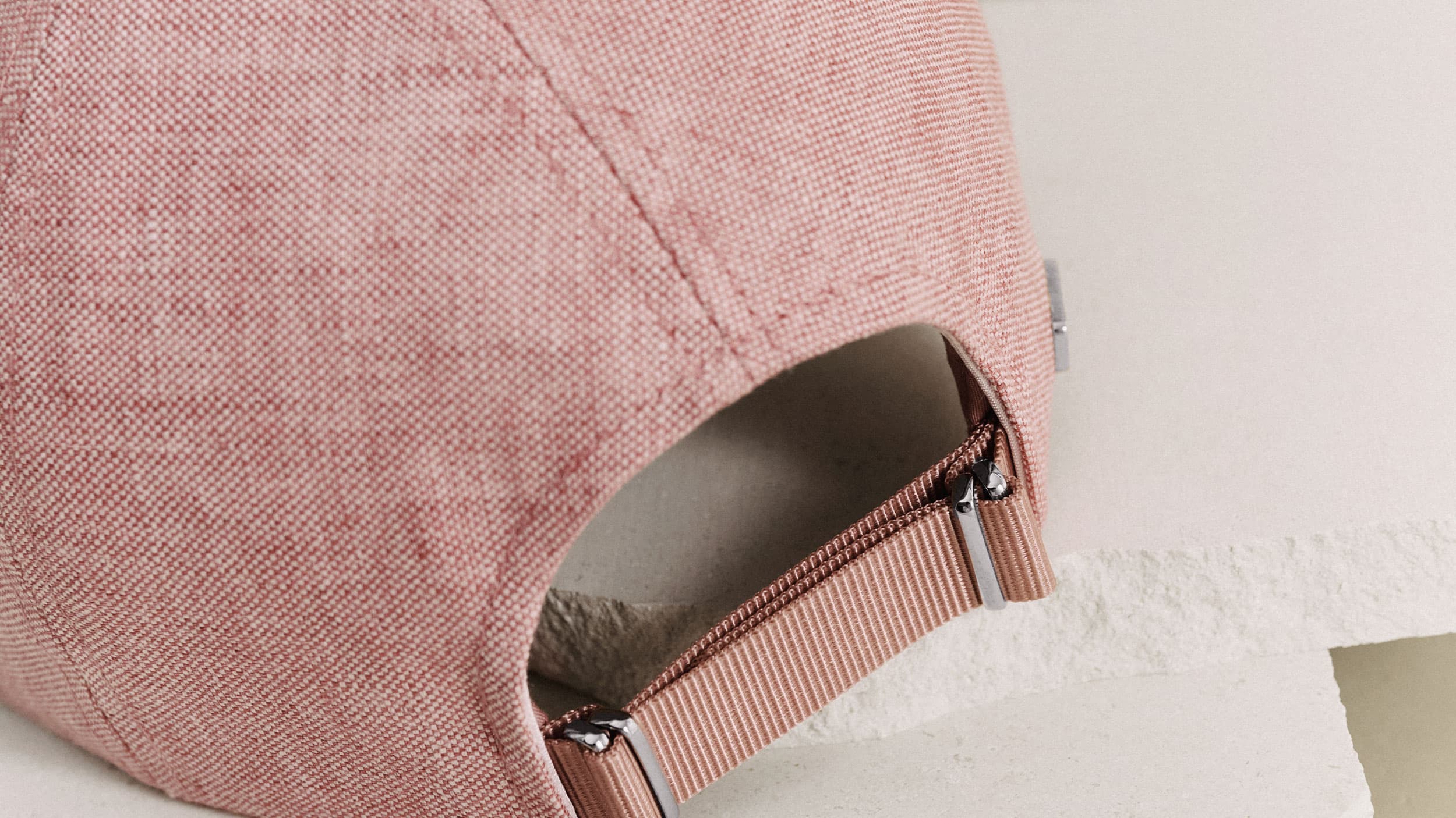 Close-up of a pink fabric cap's back, showing an adjustable strap and metal buckle detail, set on a light textured surface.