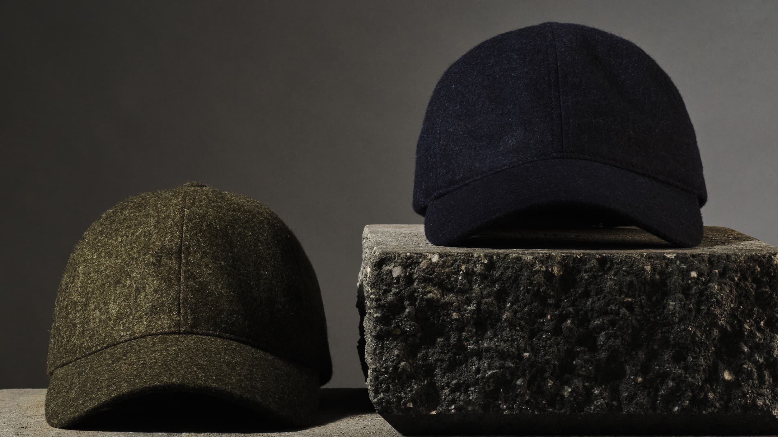 Two wool baseball caps, one olive green and one dark blue, displayed on a rough stone and concrete surface against a dark background.