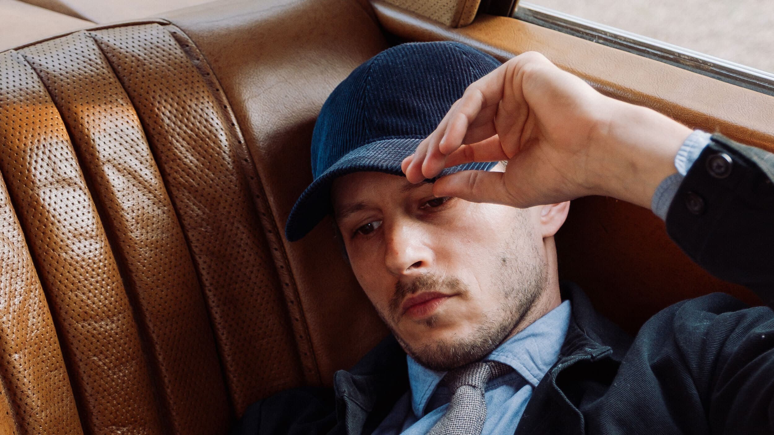Man in a blue cap and formal attire, resting against a brown leather car seat, looking contemplative.