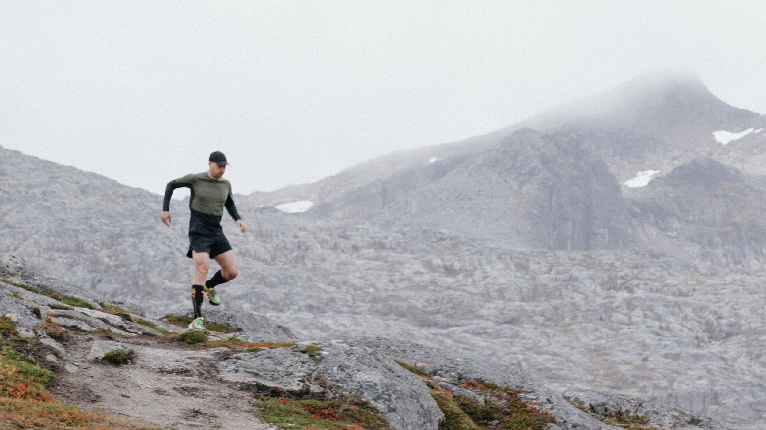 A person wearing a cap and athletic clothing is trail running on rocky terrain in a foggy mountain landscape.