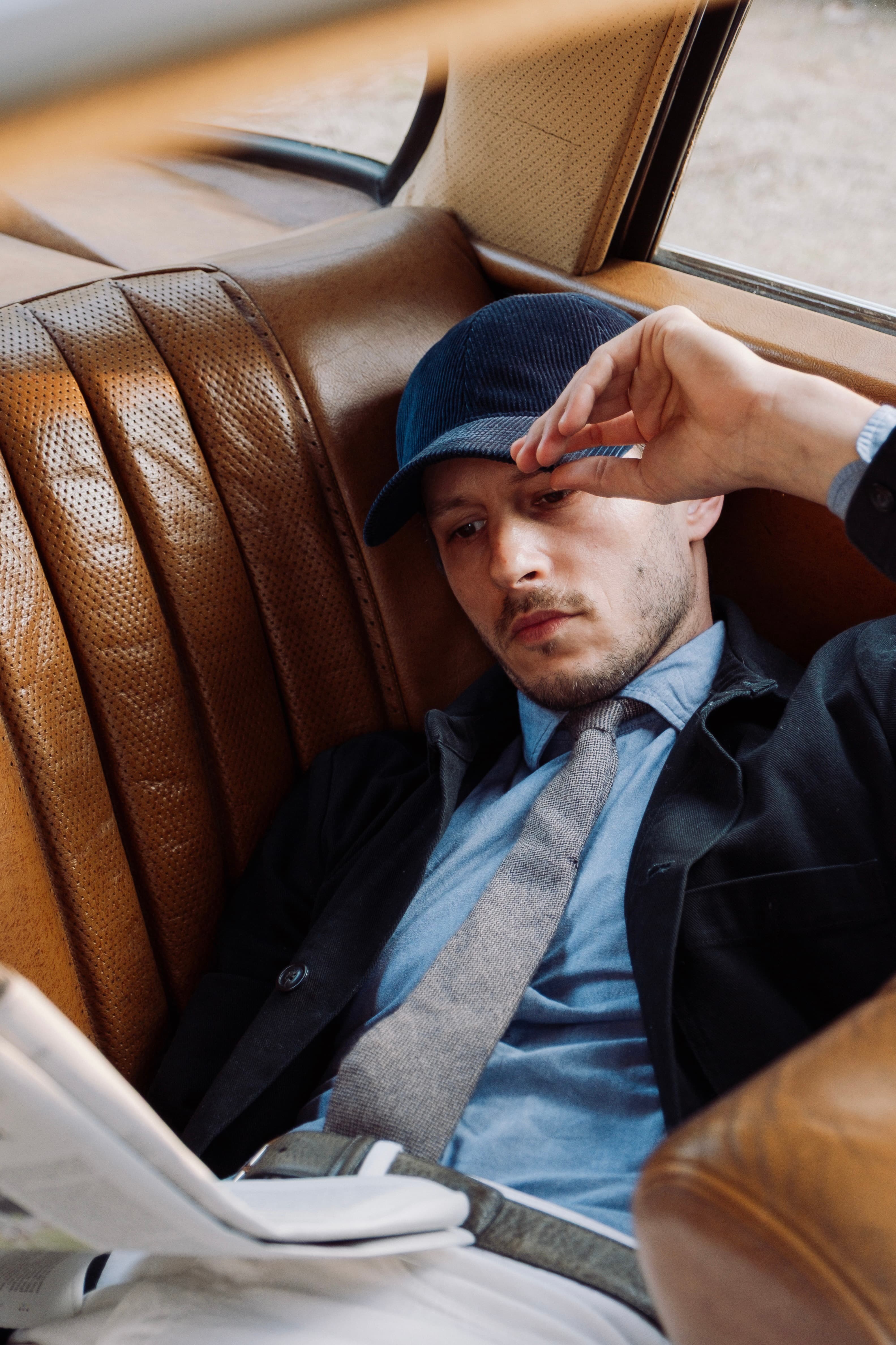 Hedi Sersoub in casual attire with a cap relaxes in the back seat of a car, reading a newspaper.