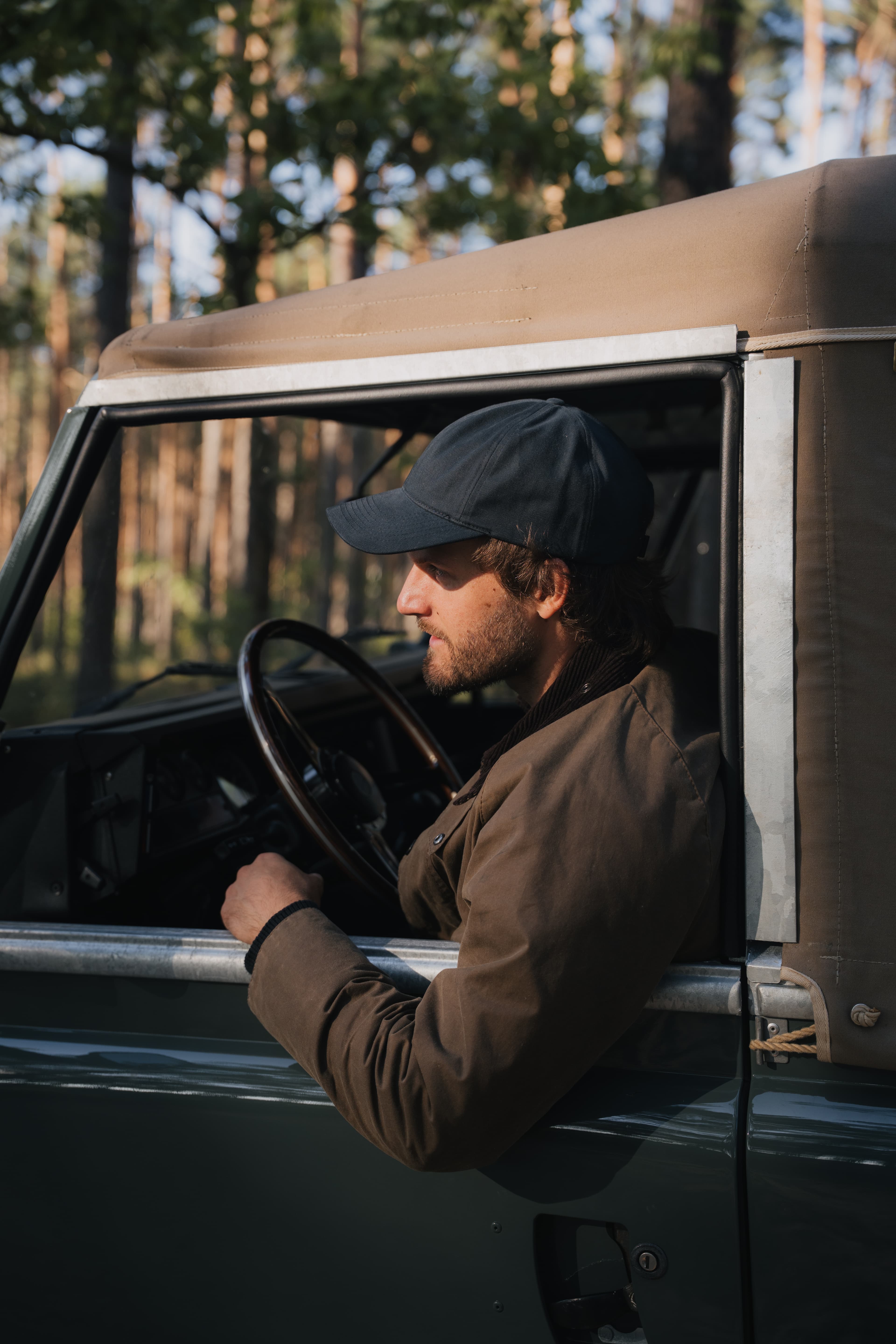 A man wearing a cap sits in an open car window, surrounded by a forest backdrop, holding the steering wheel.