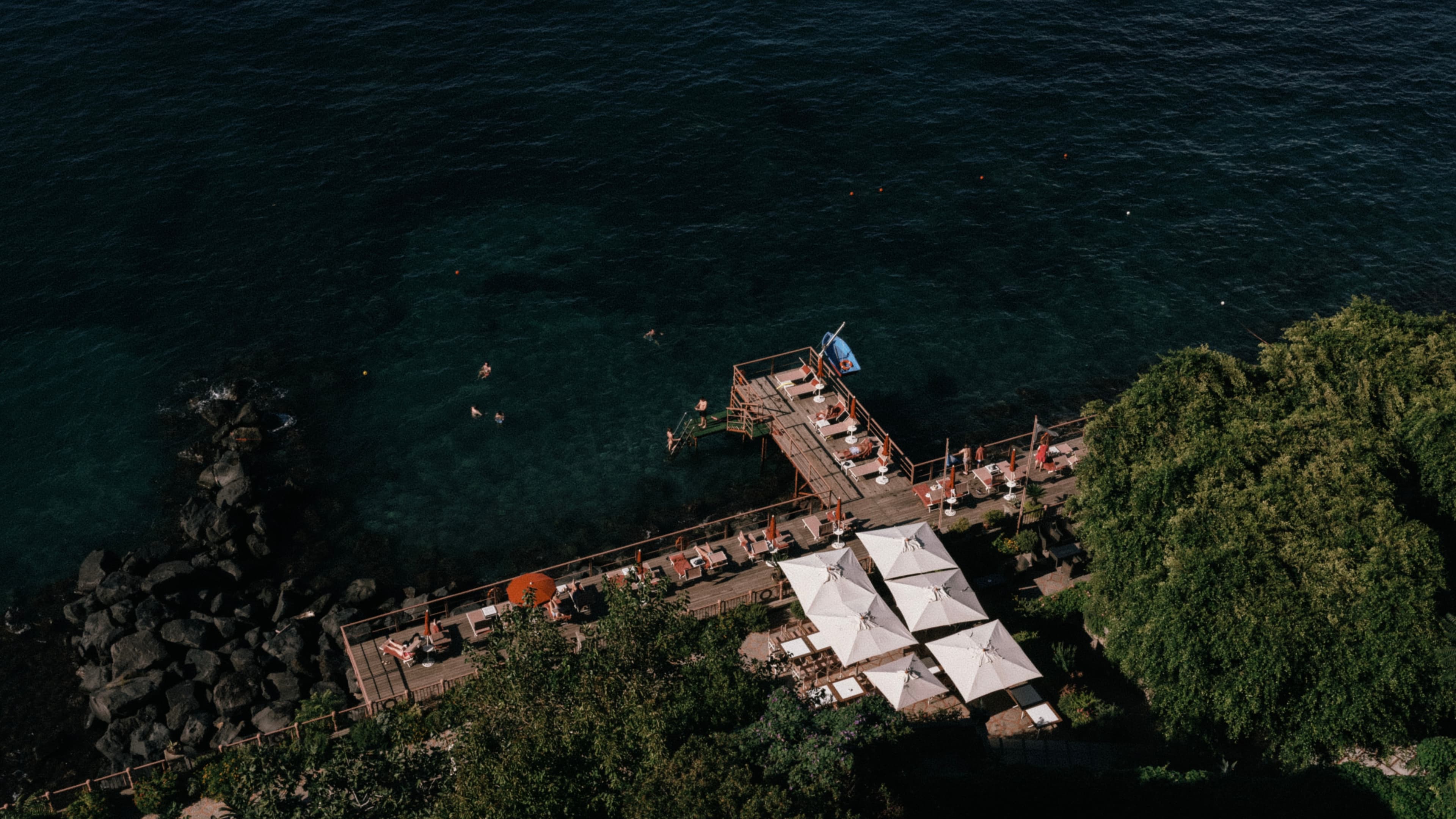 Aerial view of a dock with sun loungers, umbrellas, and people swimming in clear blue water, surrounded by lush greenery and rocky shoreline.
