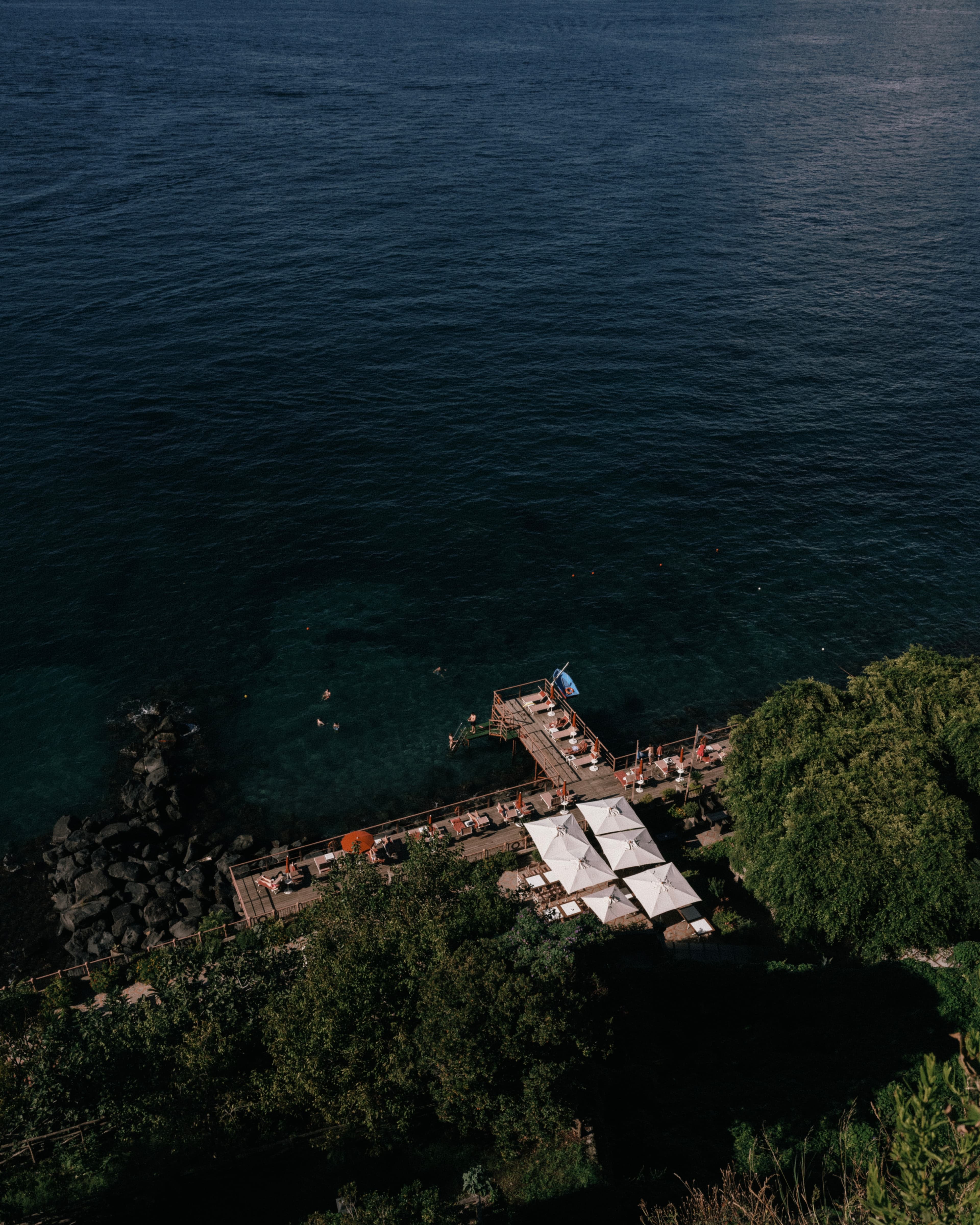 Aerial view of a beachside area with loungers and umbrellas on a dock, surrounded by trees, overlooking a calm blue sea.