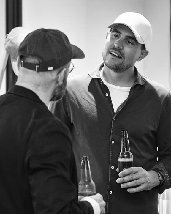 Two men in casual attire converse while holding beer bottles, both wearing caps, in a candid black-and-white photo.